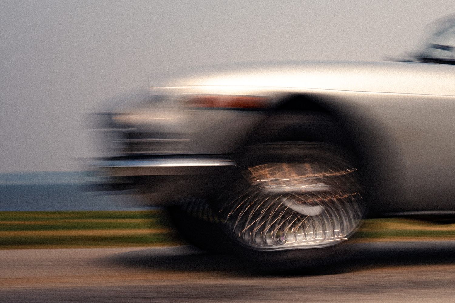 A silver classic car captured in motion blur, speeding along a road, showcasing its vintage chrome wheels.