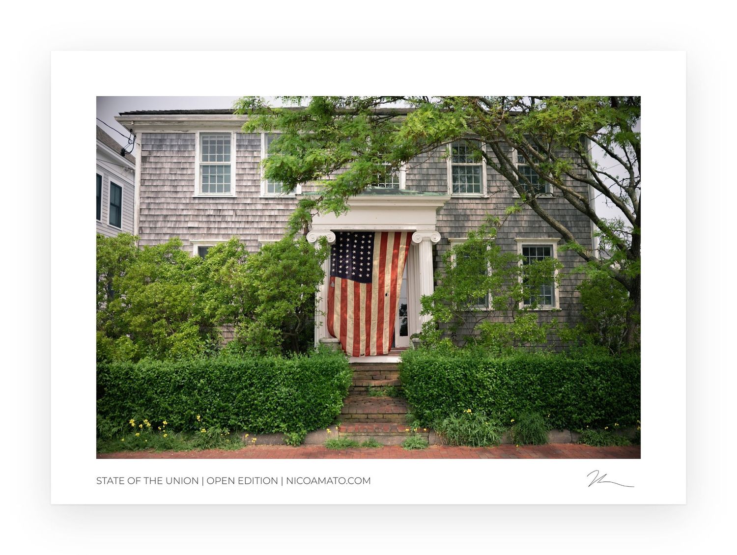 A large American flag hangs on a charming shingled home entrance surrounded by lush green trees and shrubs.