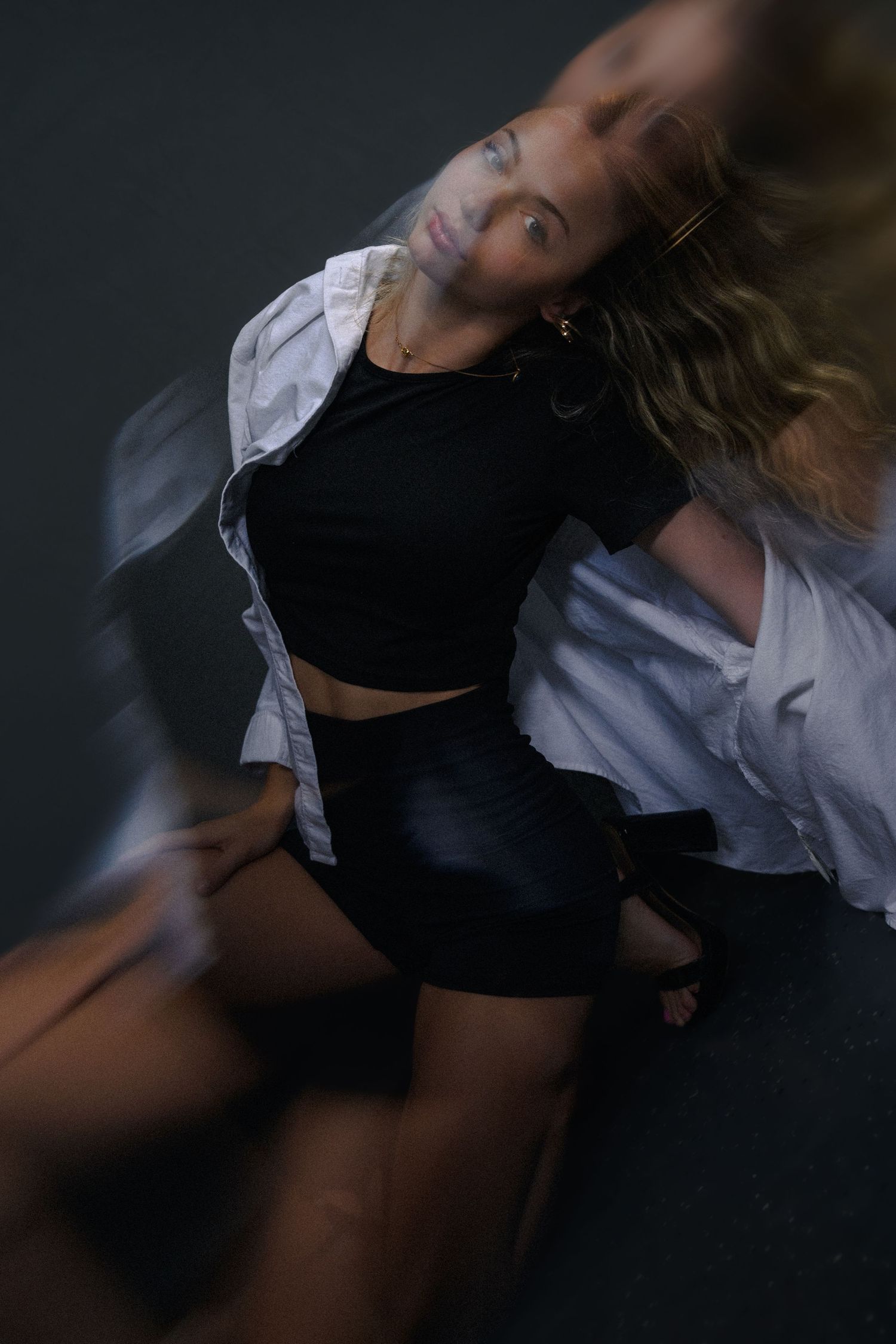 Woman in black crop top and shorts with white shirt poses dynamically with motion blur effect in dark studio.