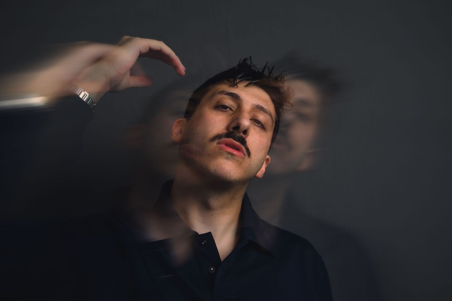 Young man with mustache in dark studio, motion blur effect creating ghostly double exposure portrait.