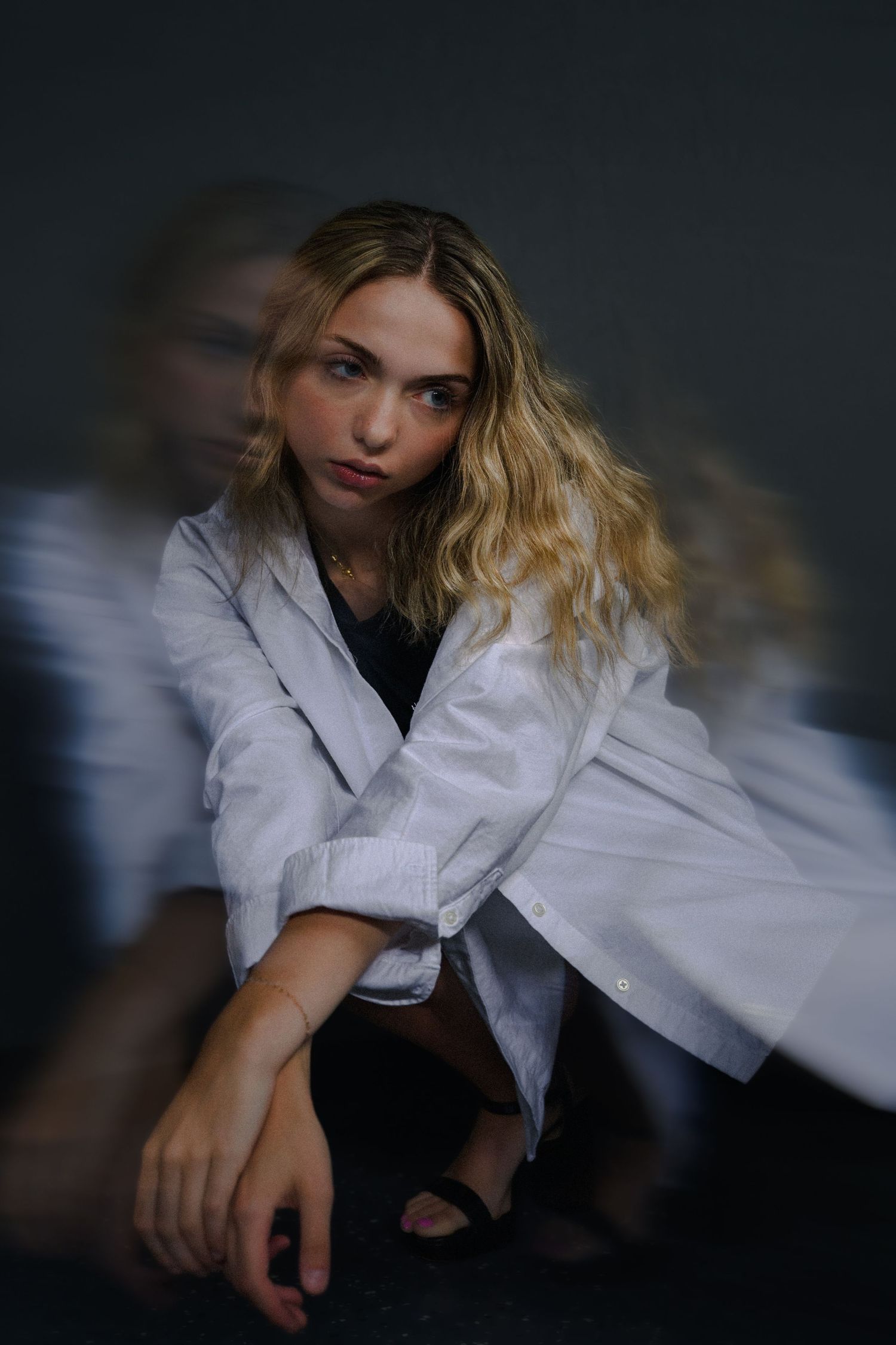 Blonde woman in white oversized shirt poses with motion blur effect against dark background.