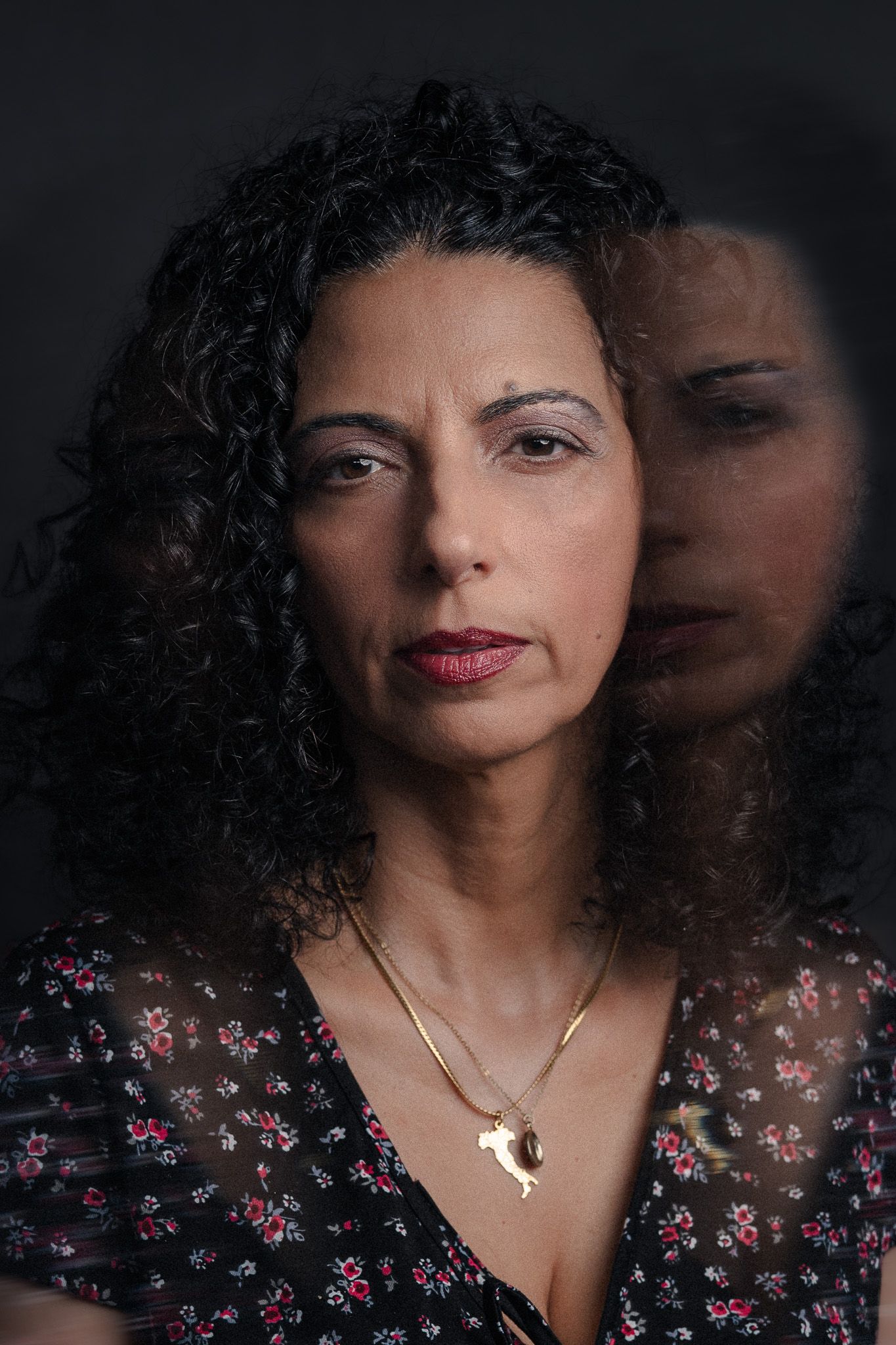 A woman with curly dark hair wearing a floral dress and gold necklace, reflected in glass against dark background.