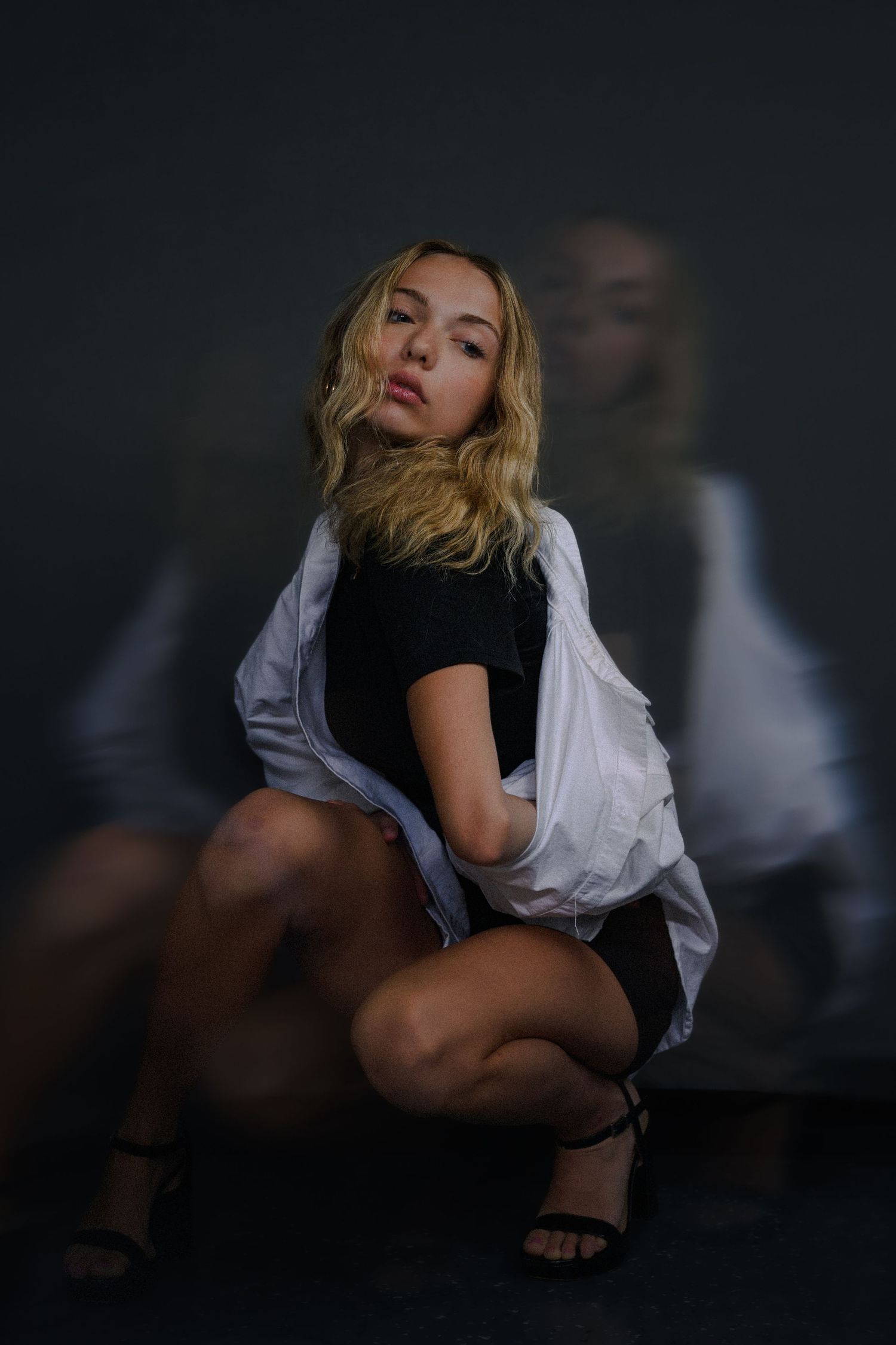 Blonde woman in black bodysuit and white shirt crouching in heels against dark reflective wall.