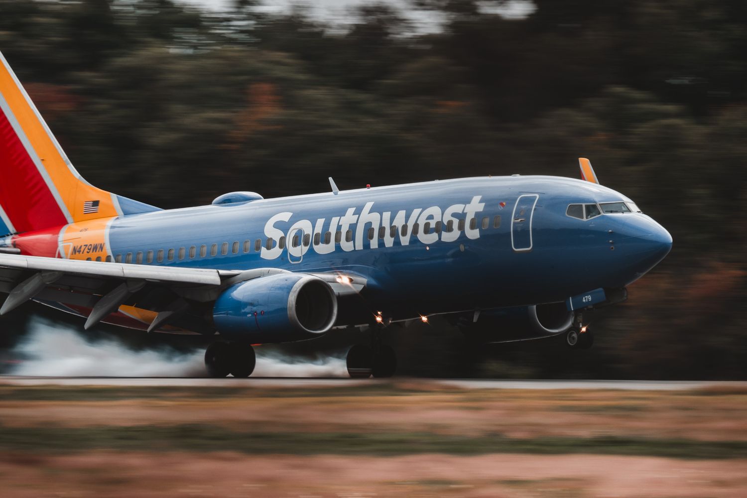 Southwest Airlines blue Boeing 737 taking off on runway with smoke from tires and blurred trees in background.