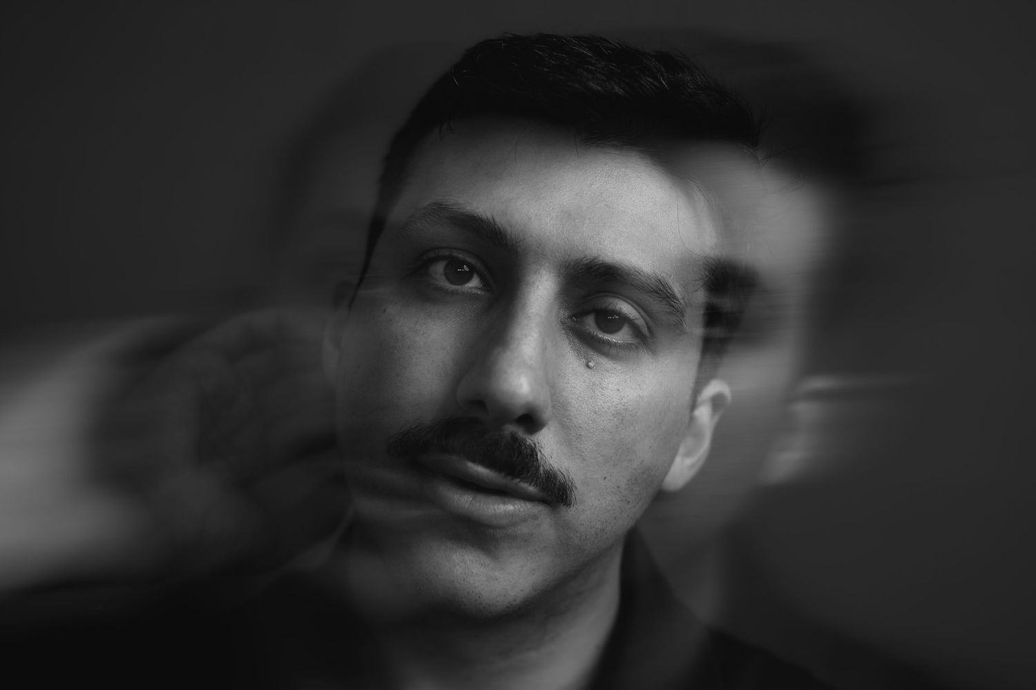 Black and white portrait of a young man with a mustache, featuring a blurred double exposure artistic effect.