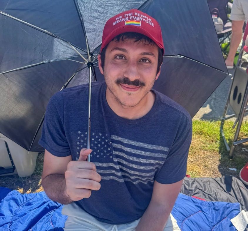 Young man with mustache sitting outdoors under black umbrella wearing red cap and American flag t-shirt.