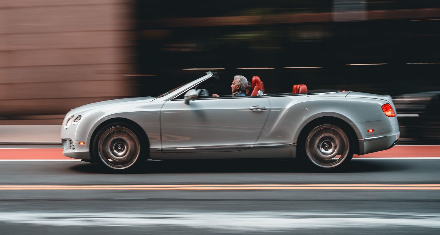 Silver Bentley Continental GT convertible speeding through city street with red interior and bronze wheels.