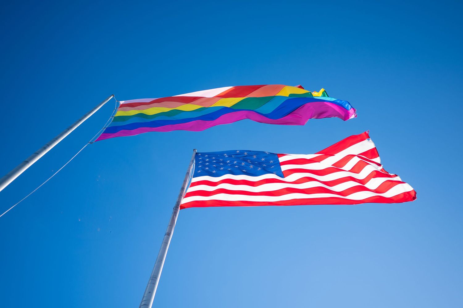 Rainbow Pride flag and American flag flying together against a clear blue sky.