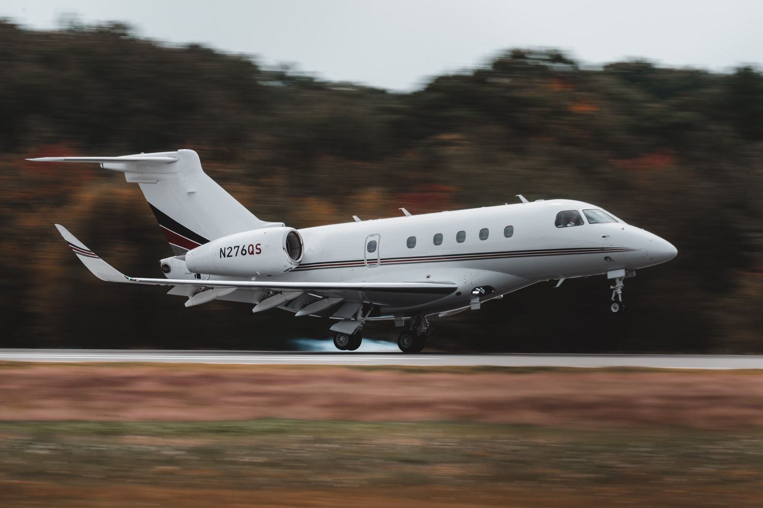 White private jet N274QS taking off on runway with blurred autumn trees in background.