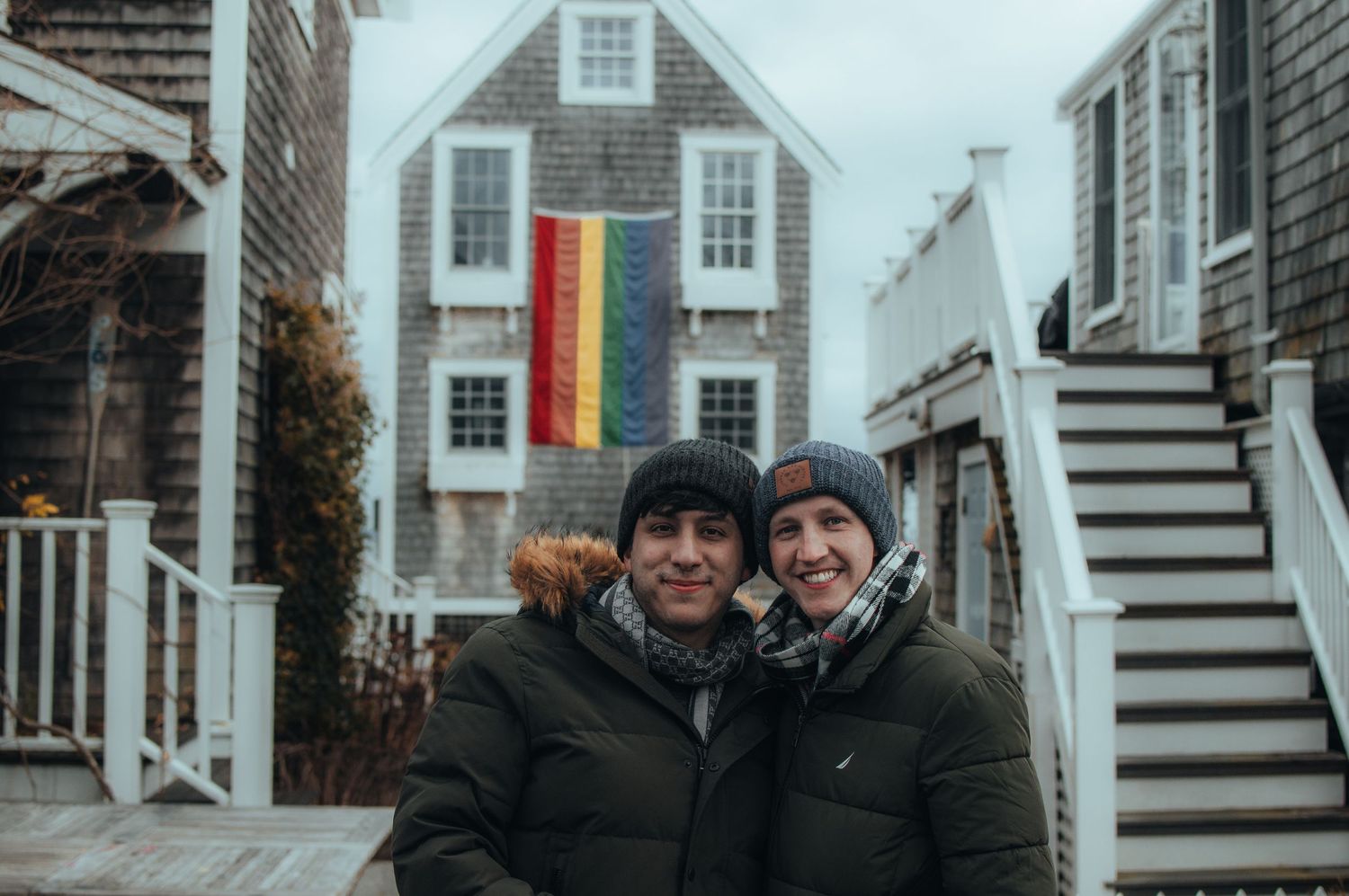 Couple smiling together in front of a house displaying a rainbow Pride flag.