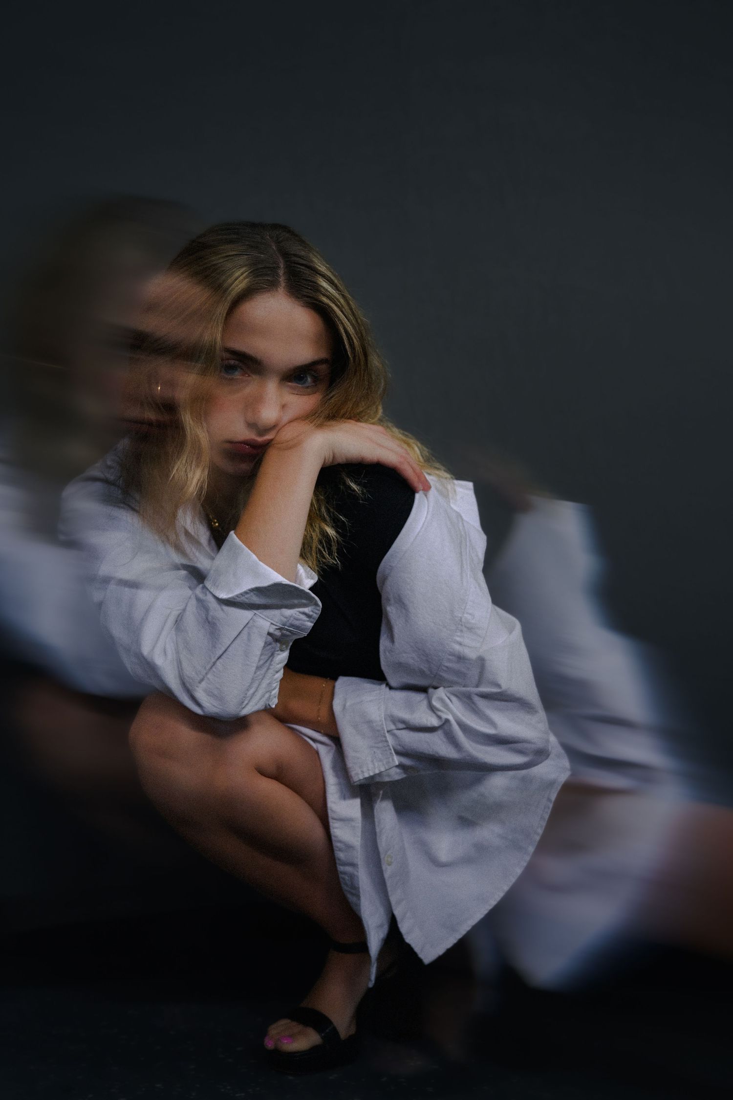 Young blonde woman in white oversized shirt sitting pensively, with artistic motion blur effect creating ghostly double exposure.