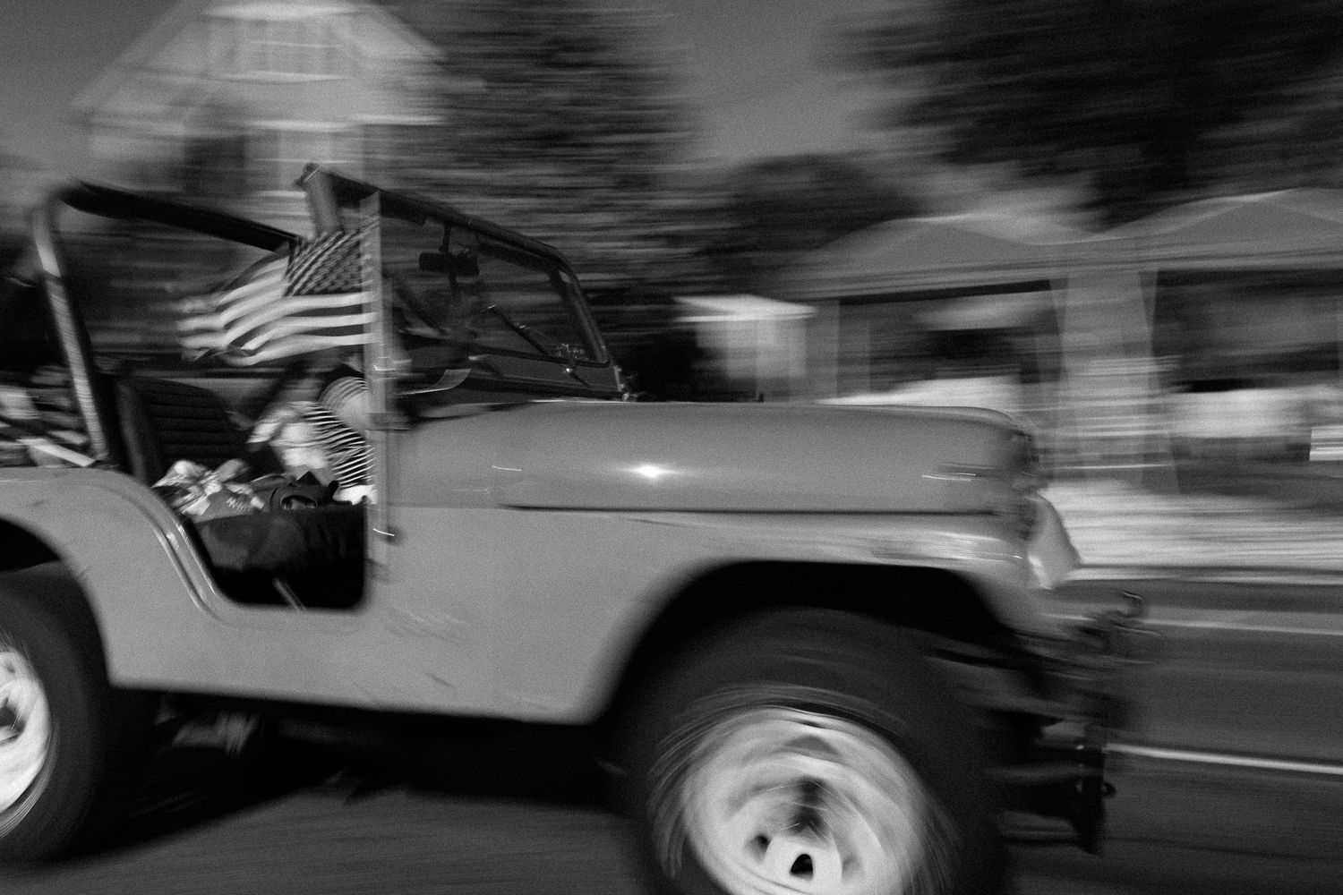 Black and white photo of a jeep carrying an American flag driving in a parade, captured with motion blur.