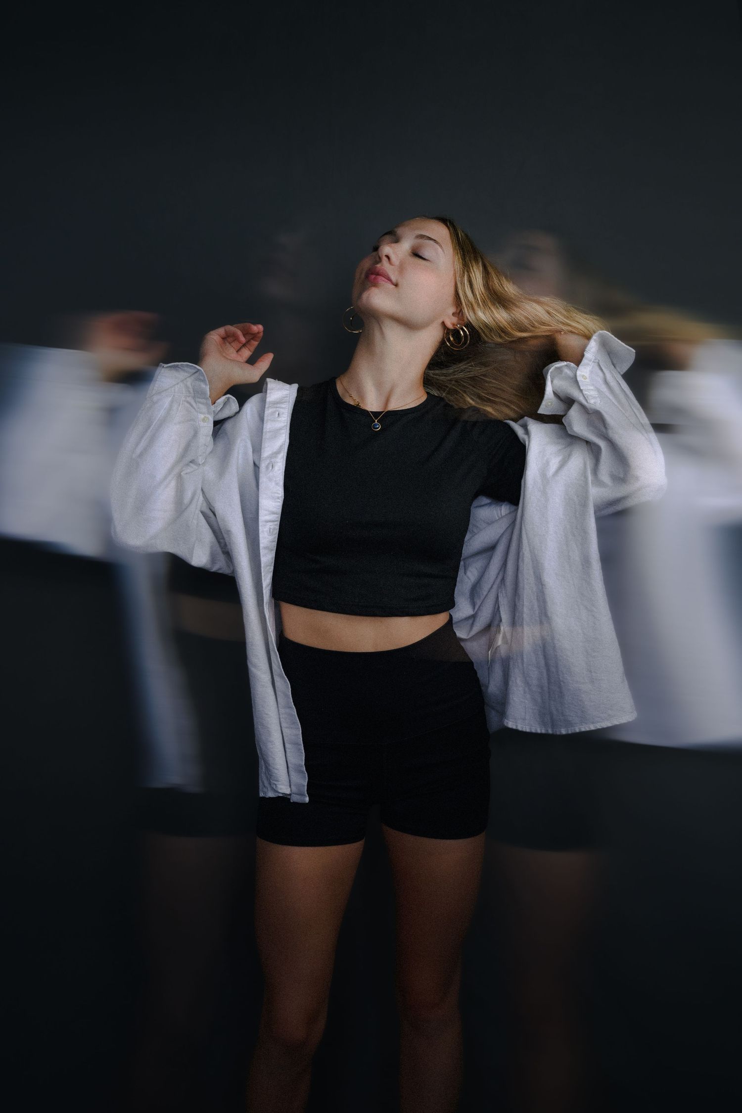 Young woman in black crop top and shorts with open white shirt, posing dynamically with motion blur effect on dark background.