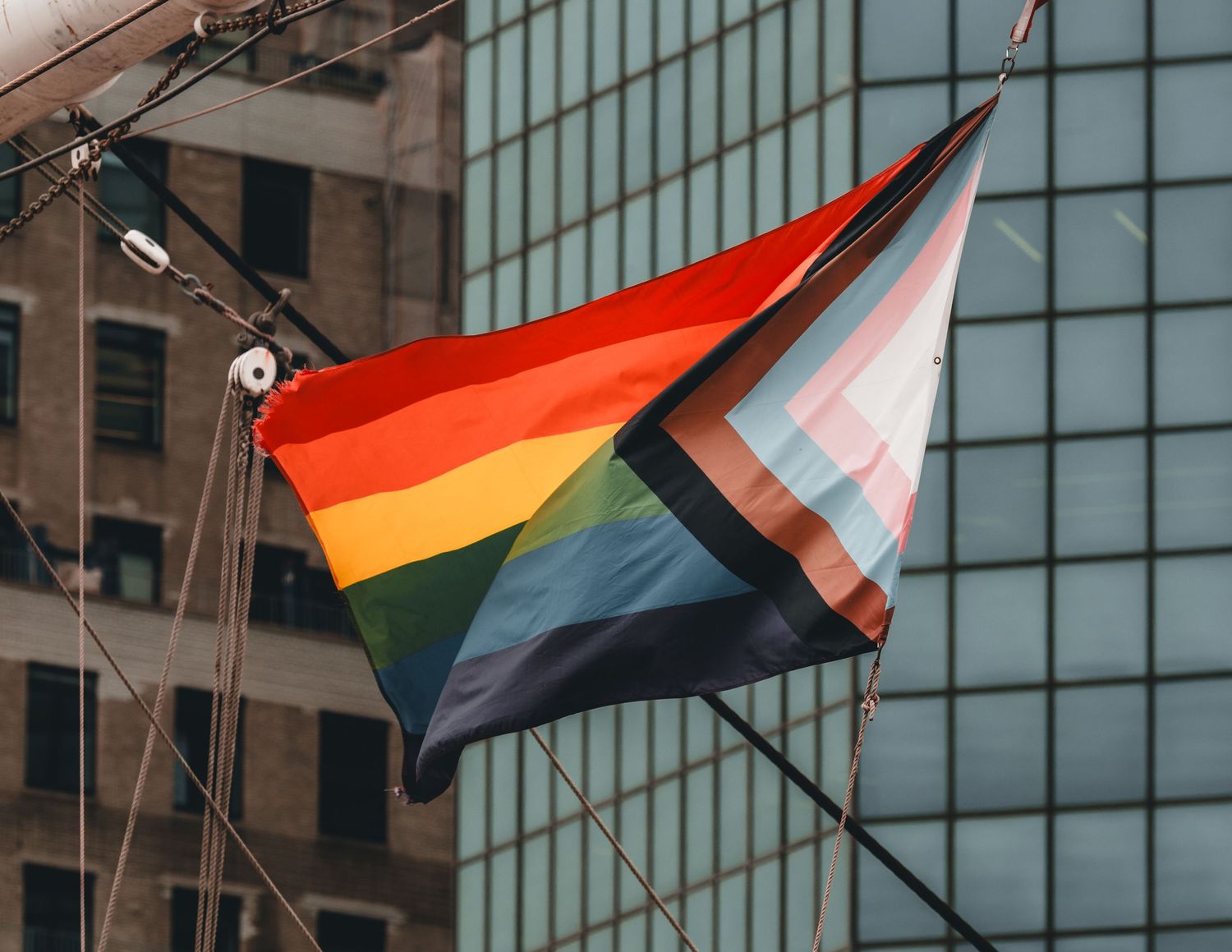 Progress Pride flag flying from a mast against a glass building in a city.