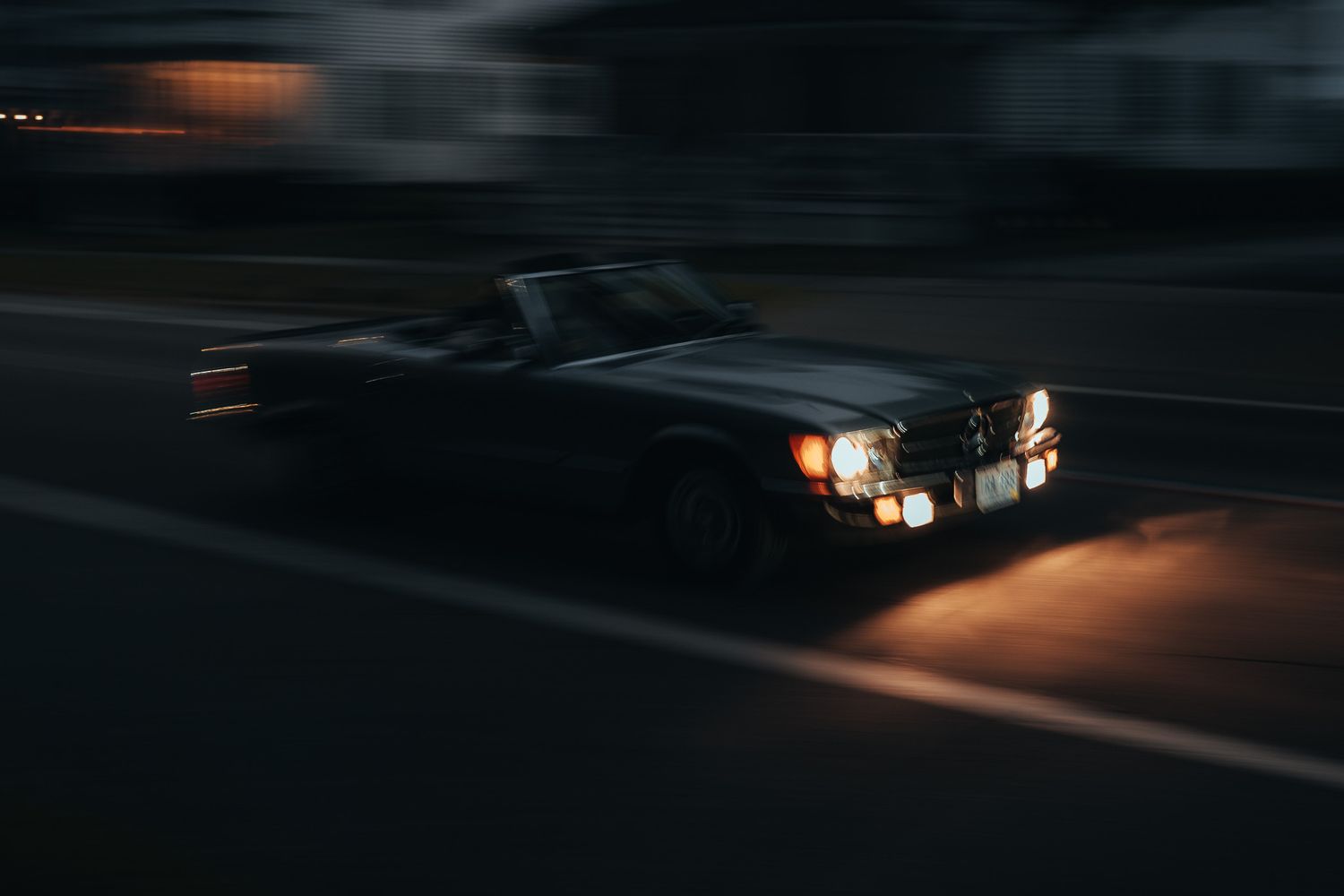 A classic vintage convertible car driving at night with headlights on, captured with motion blur effect.