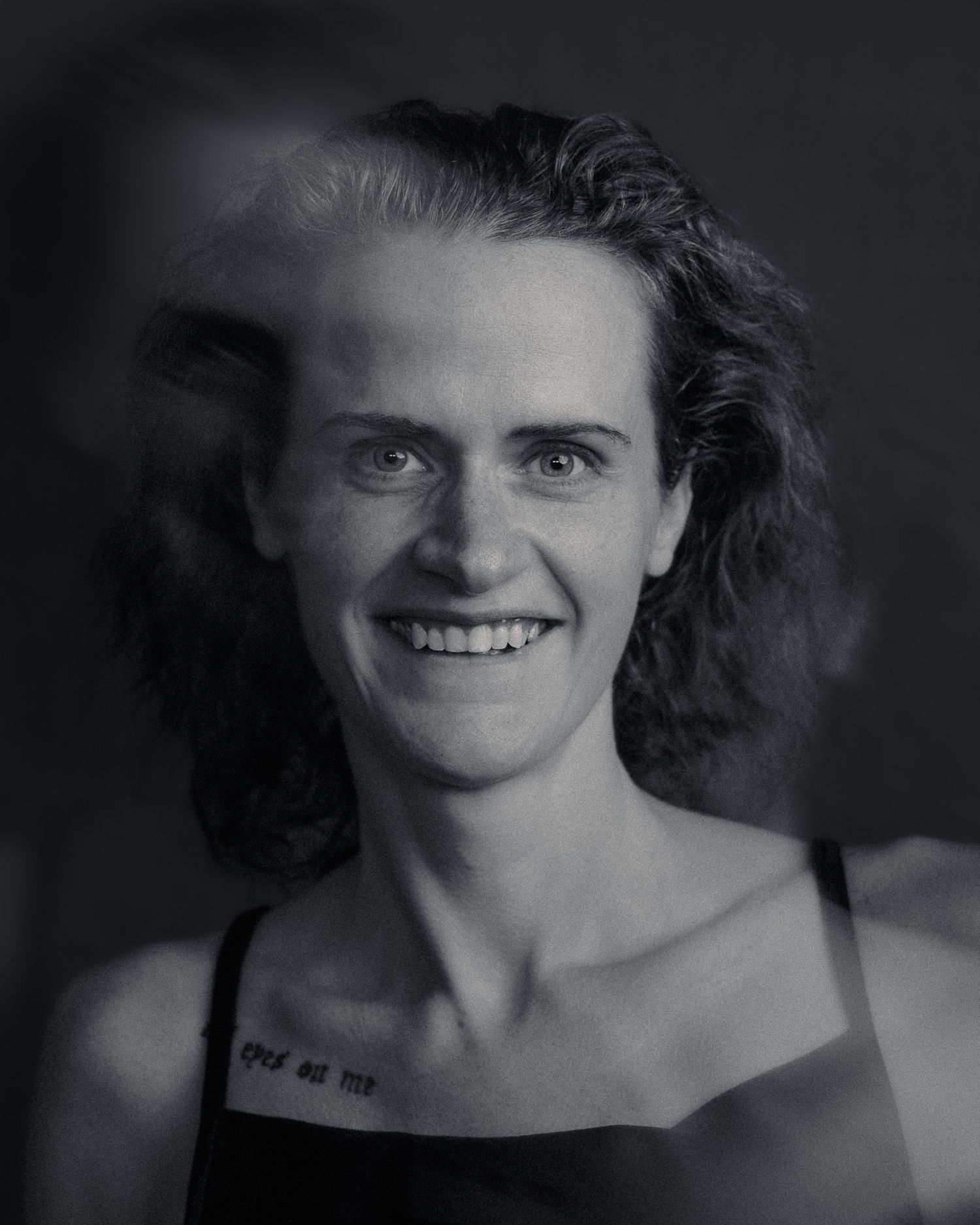 Black and white portrait of a smiling woman with curly hair and a tattoo on her chest.