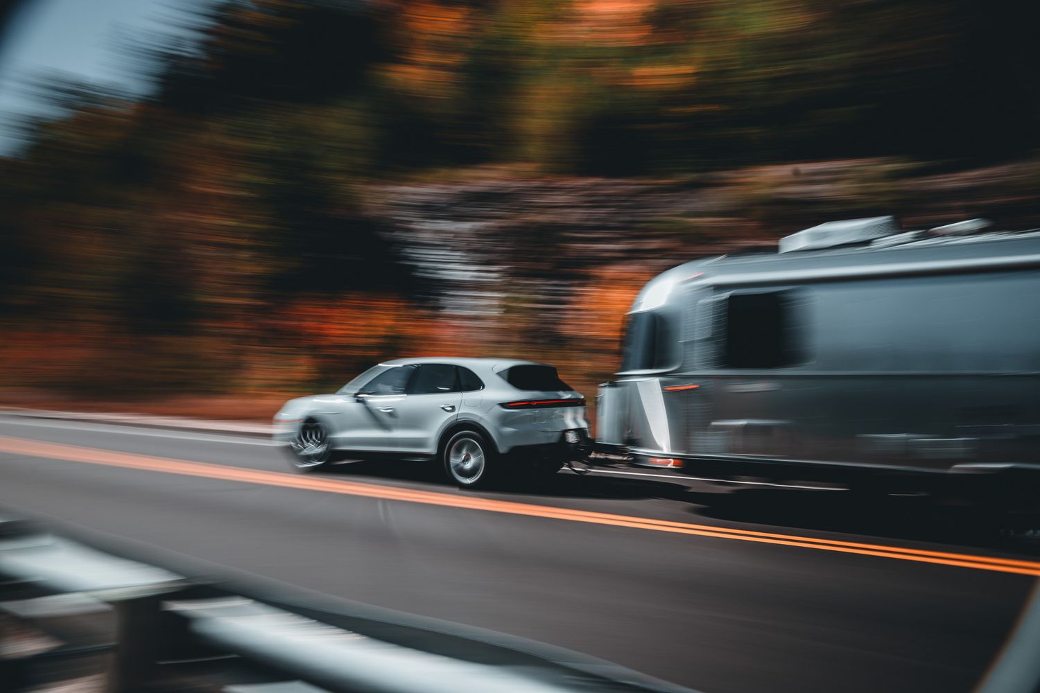 Silver SUV towing an Airstream trailer at high speed on a scenic highway with motion blur effect.