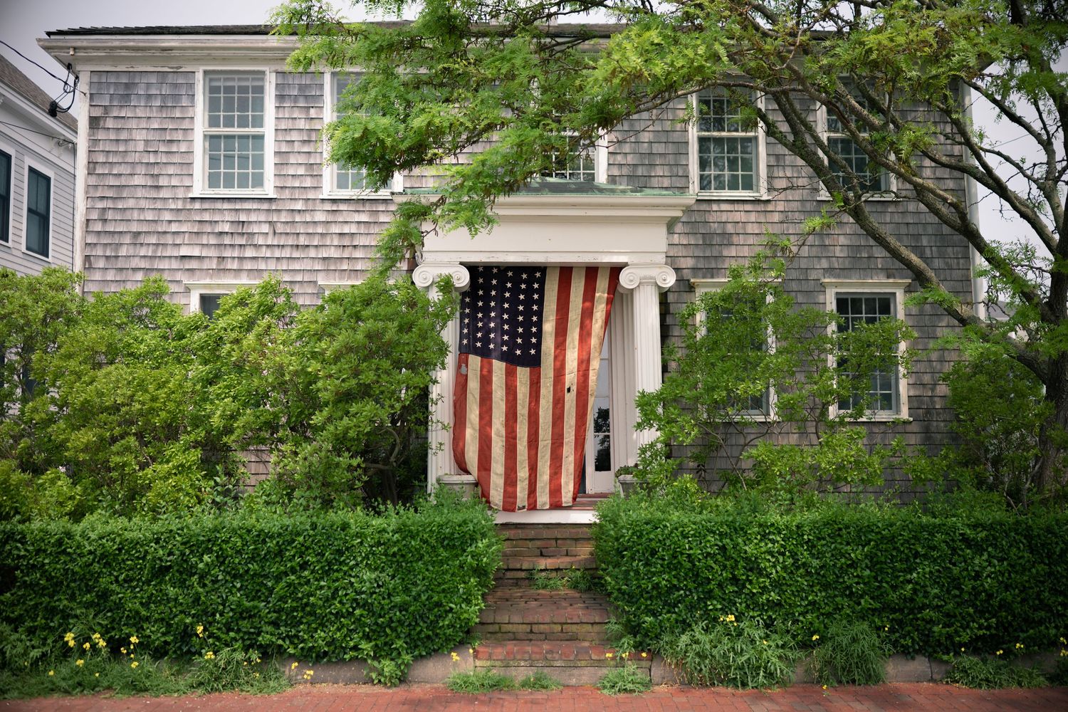 Charming gray shingle colonial home with American flag on front door, surrounded by lush green hedges and trees.