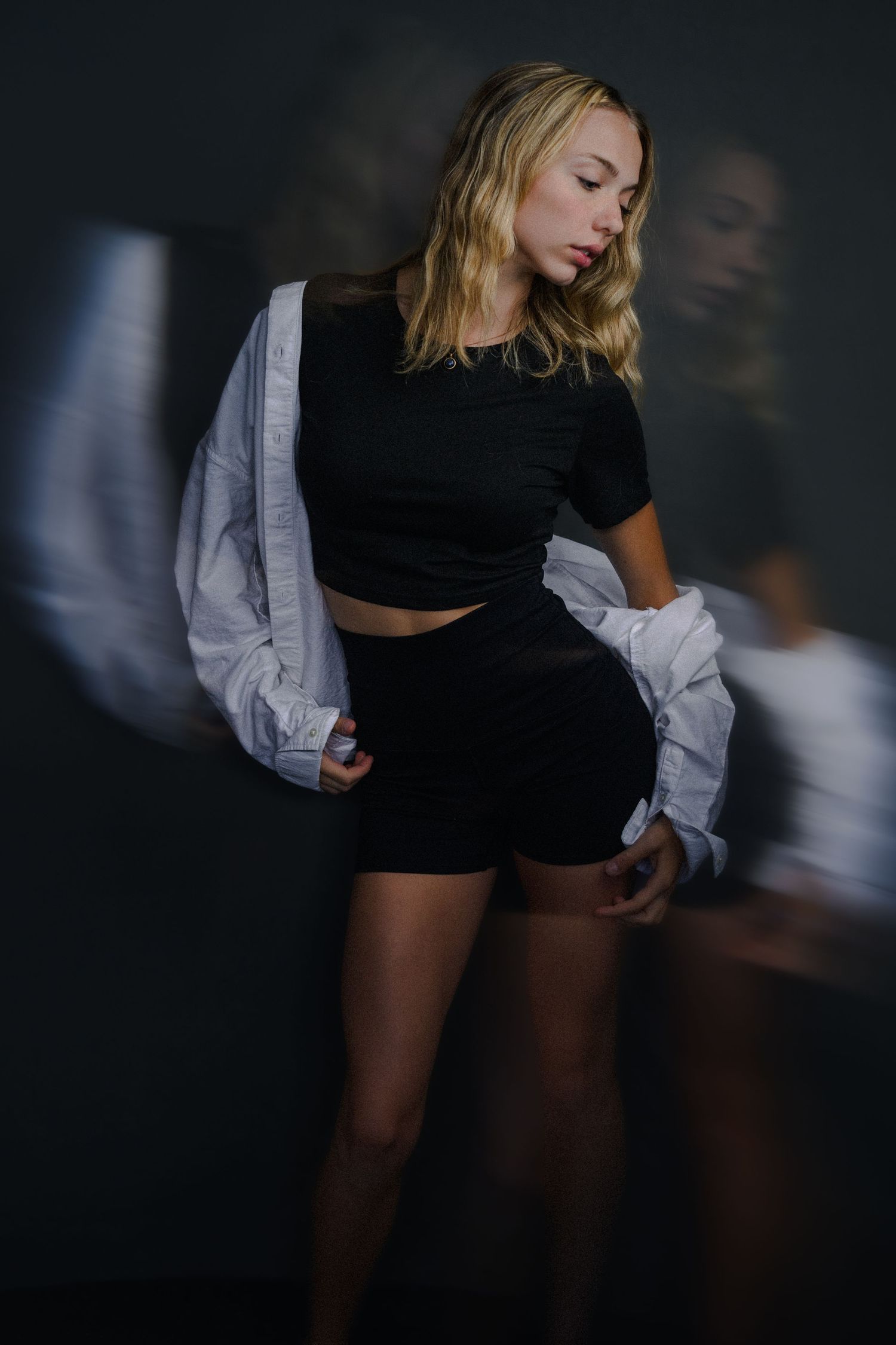 Blonde woman in black crop top and shorts with white jacket posed against dark background.