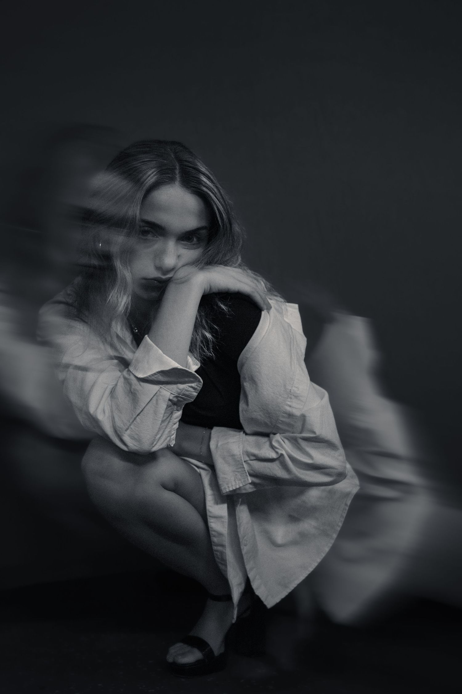 Black and white long exposure portrait of a woman in a white blouse crouching thoughtfully against a dark background.