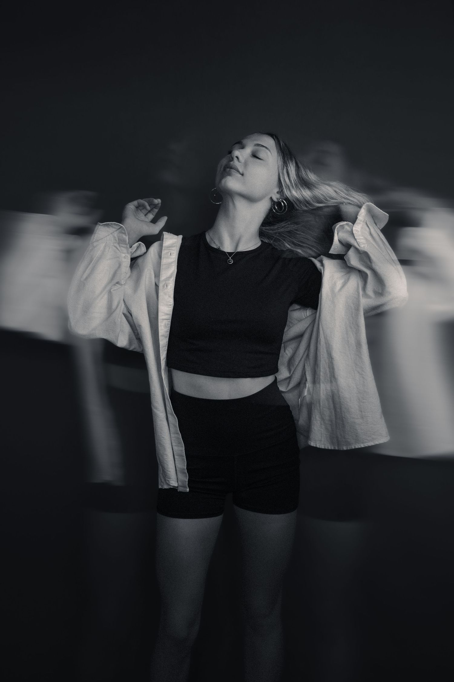 Black and white long exposure photo of a young woman dancing in a crop top, shorts, and open jacket.
