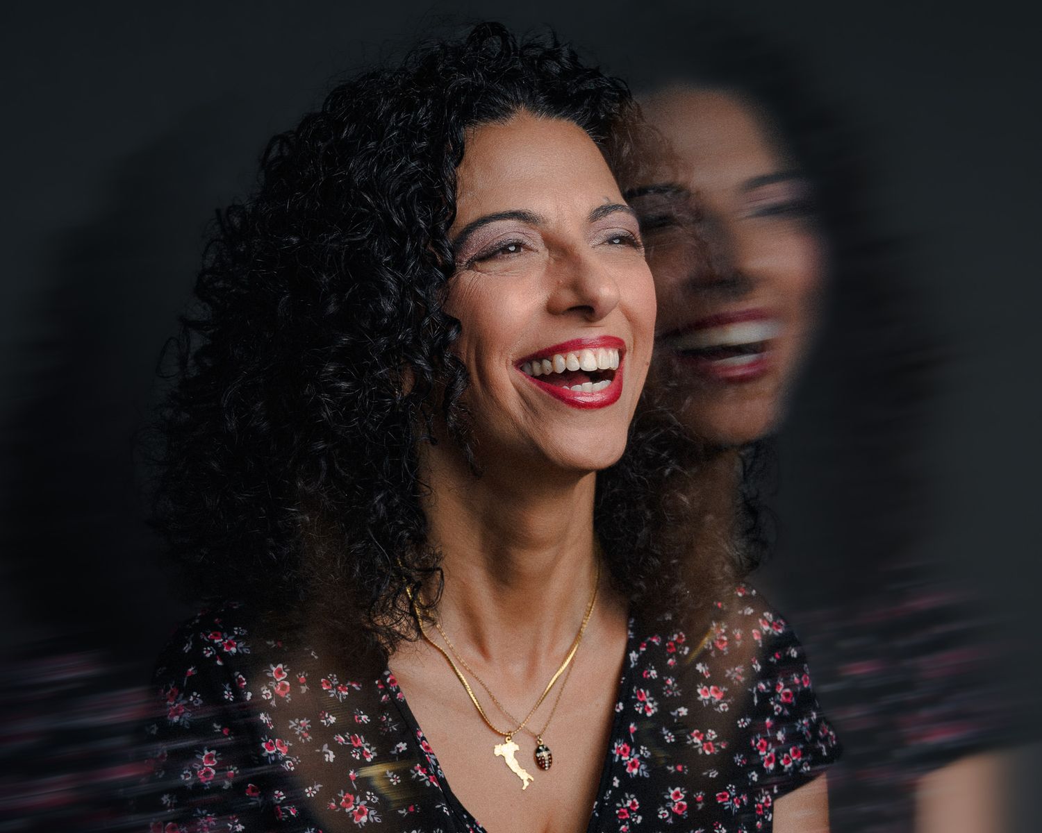 A smiling woman with curly hair wearing a floral dress and gold necklaces, laughing joyfully against a dark background.