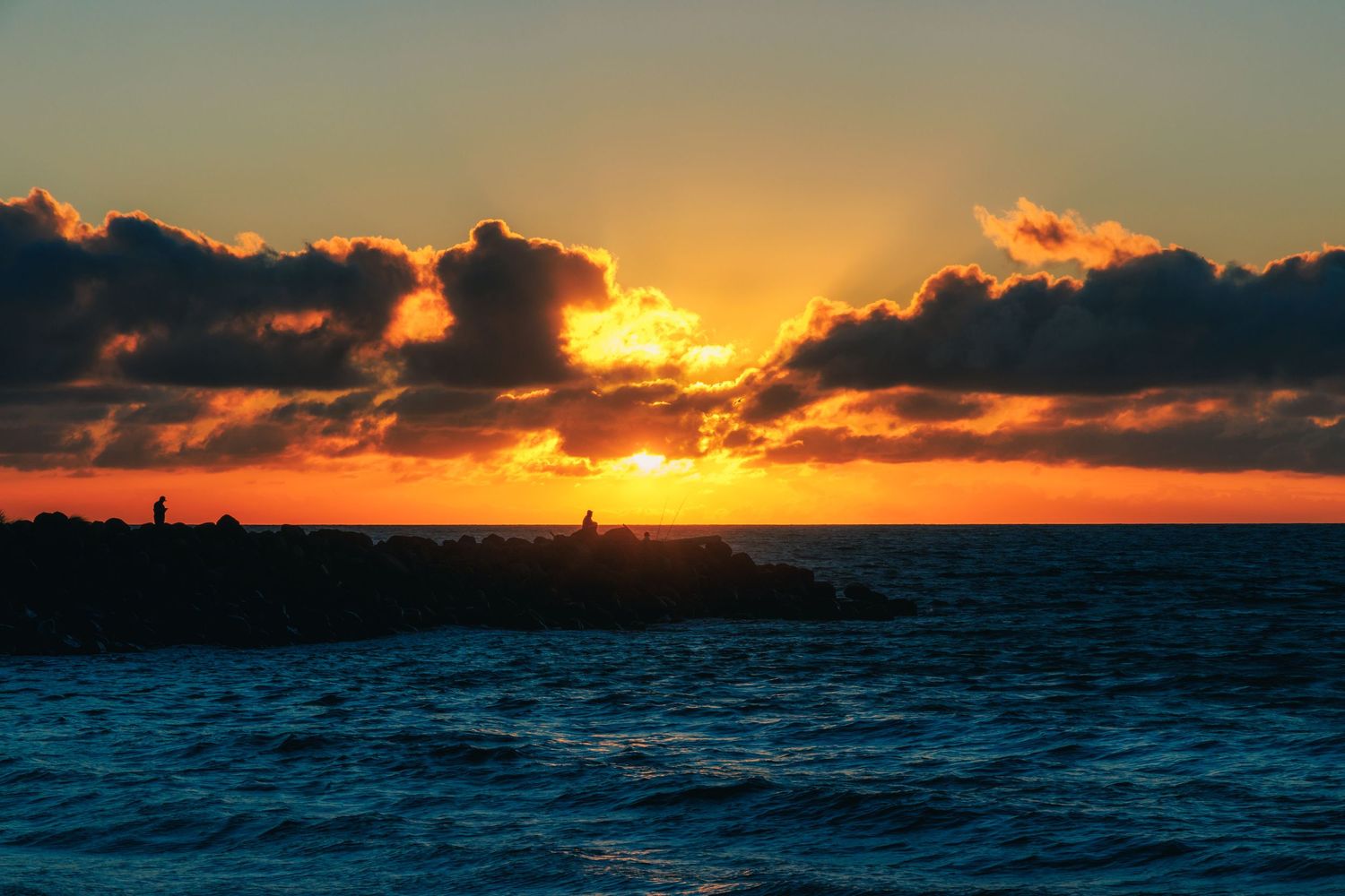 Golden sunset breaking through dramatic clouds over the Taranaki coast, with two people silhouetted on the rocks near New Plymouth, New Zeal