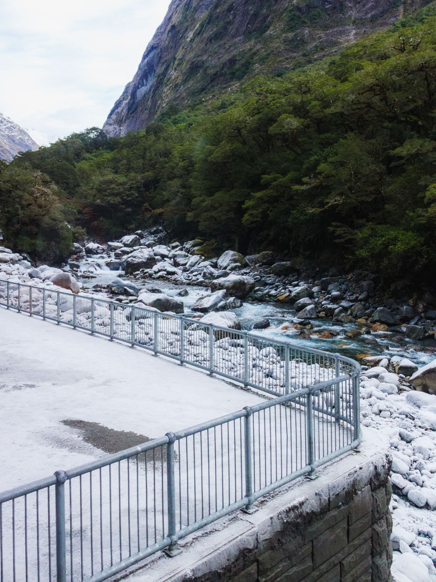 Snow-covered viewing platform beside a rocky river beneath steep forested mountains in Fiordland, New Zealand.