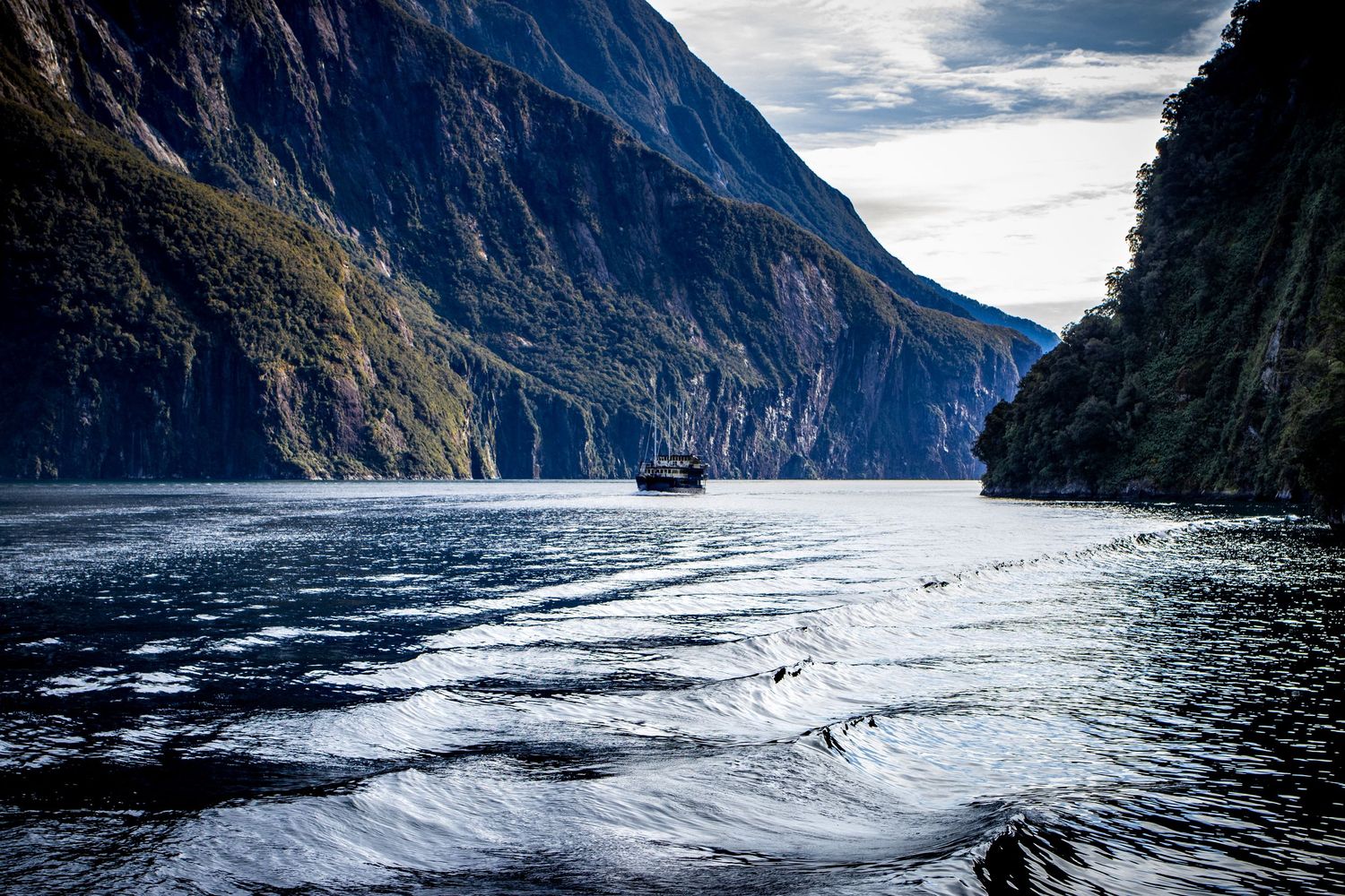 A small boat cruises through Milford Sound, surrounded by towering mountains and calm waters in Fiordland, New Zealand.