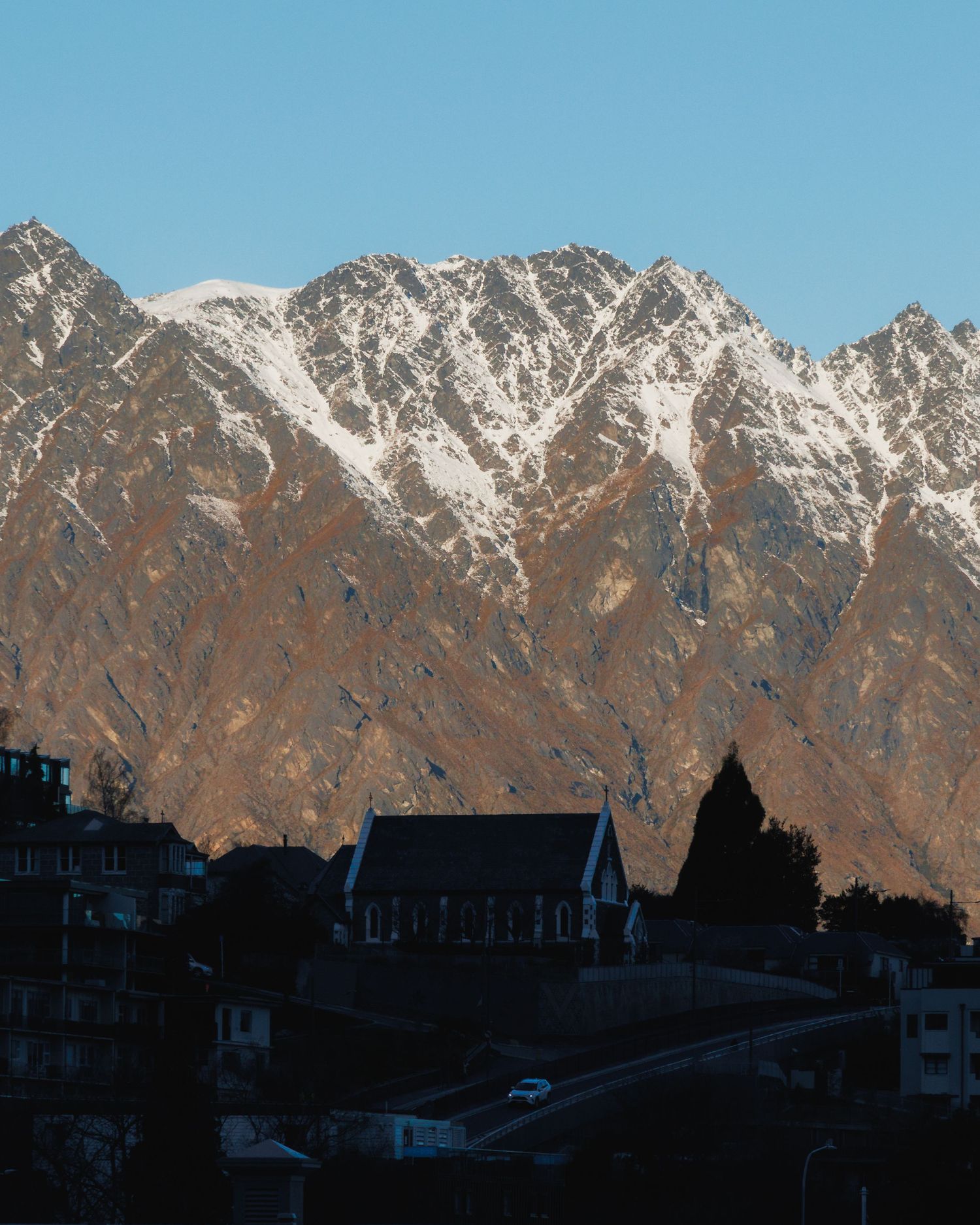 A Queenstown church and hillside homes beneath the snow-covered Remarkables on a clear winter afternoon.