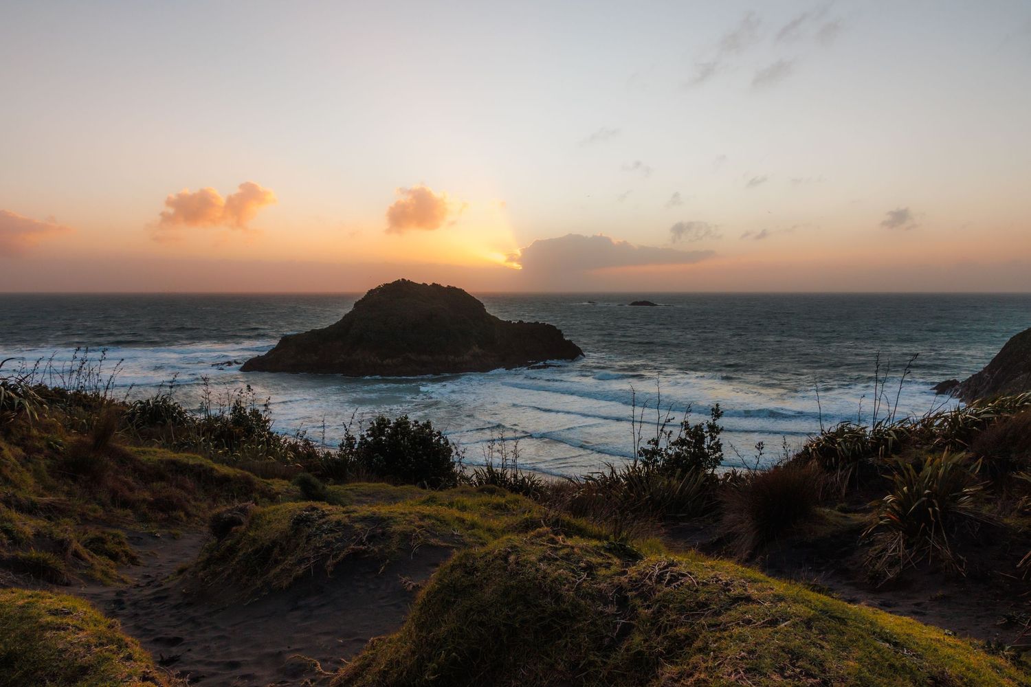 Sunset over Back Beach in New Plymouth, with dark coastal vegetation, an offshore island and waves beneath a warm glowing sky.