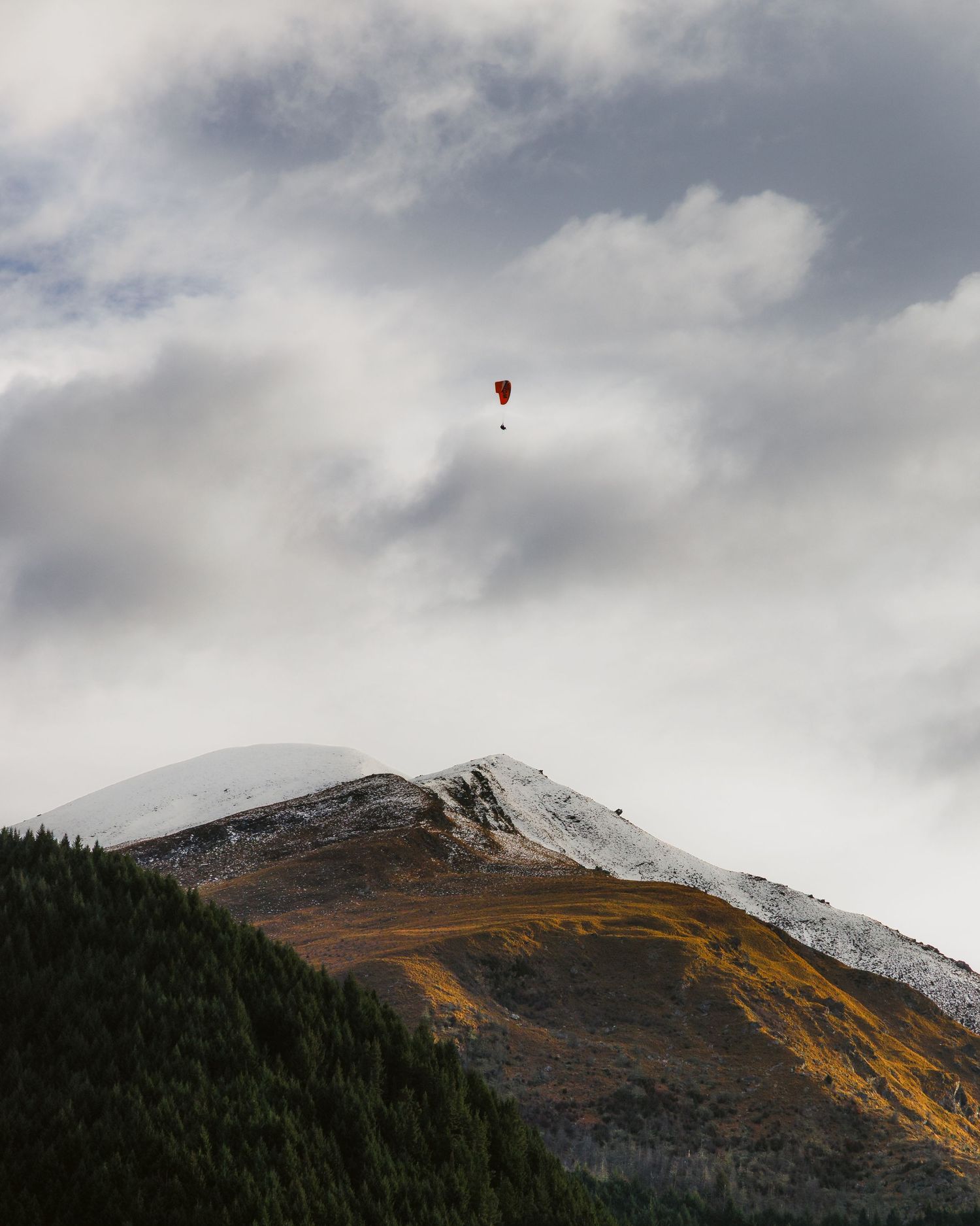 A paraglider floating above snow-dusted mountains near Queenstown, New Zealand, beneath a dramatic cloudy sky.
