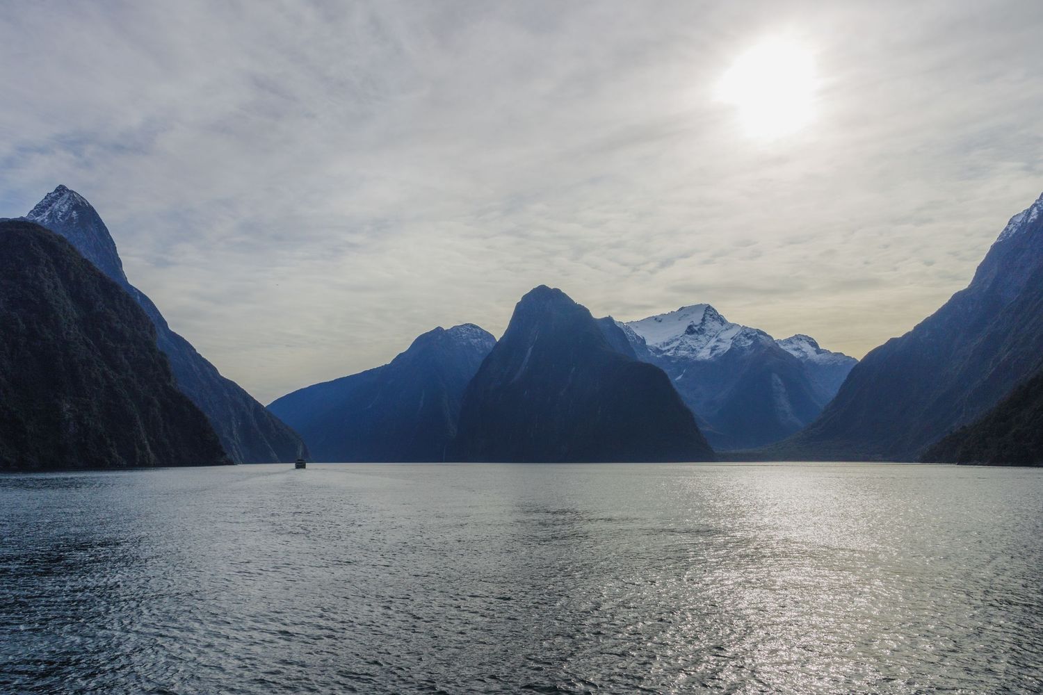 A small boat crosses Milford Sound beneath towering mountains and a bright hazy sky in Fiordland, New Zealand.