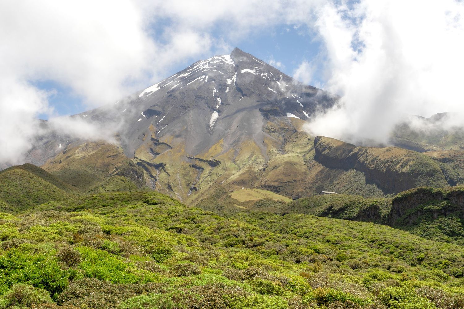 Mount Taranaki stands above green hills with patches of lingering snow beneath drifting clouds in Taranaki, New Zealand.