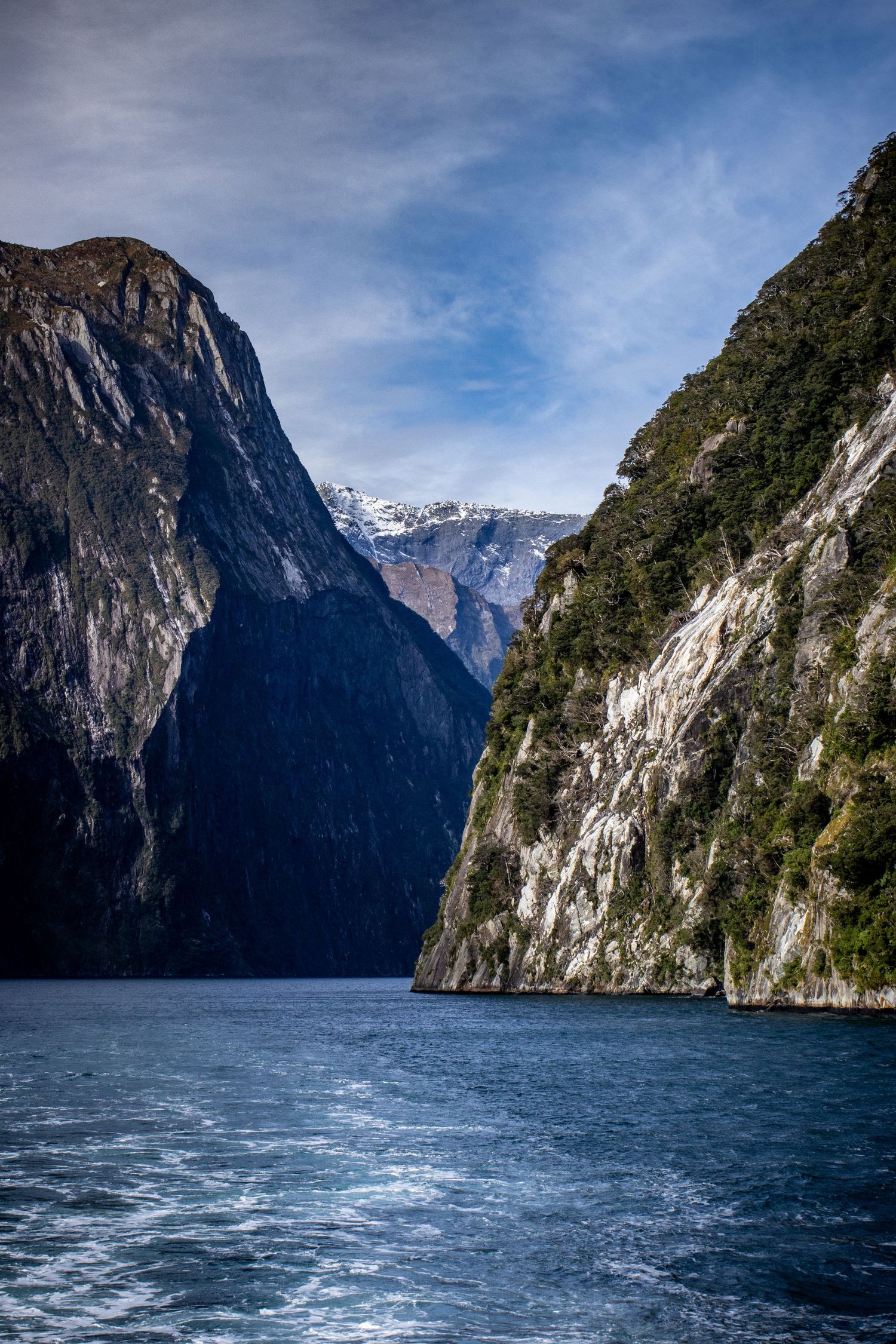 Towering mountain cliffs surround the waters of Milford Sound in Fiordland, New Zealand, beneath a partly cloudy sky.