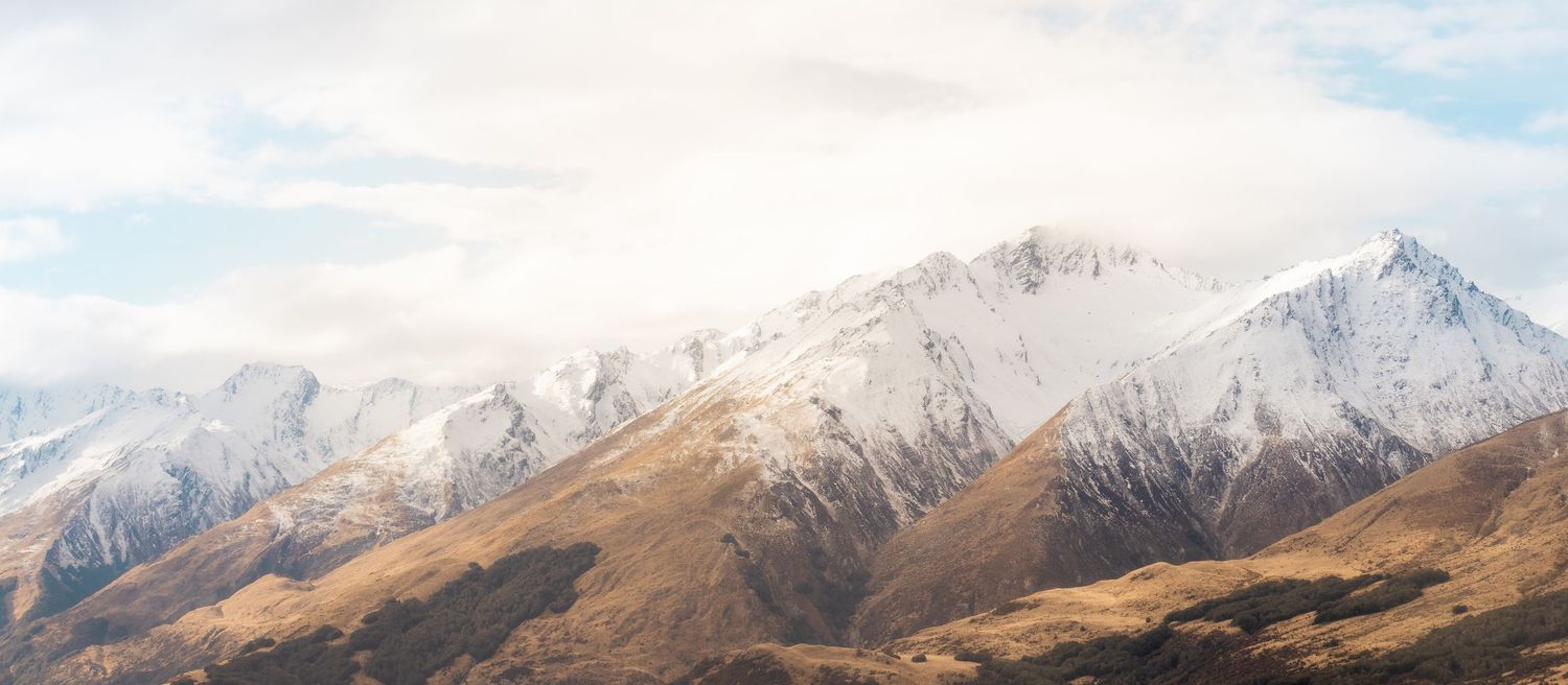 Snow-capped mountains rise above golden grasslands near Glenorchy, New Zealand, beneath a partly cloudy sky.