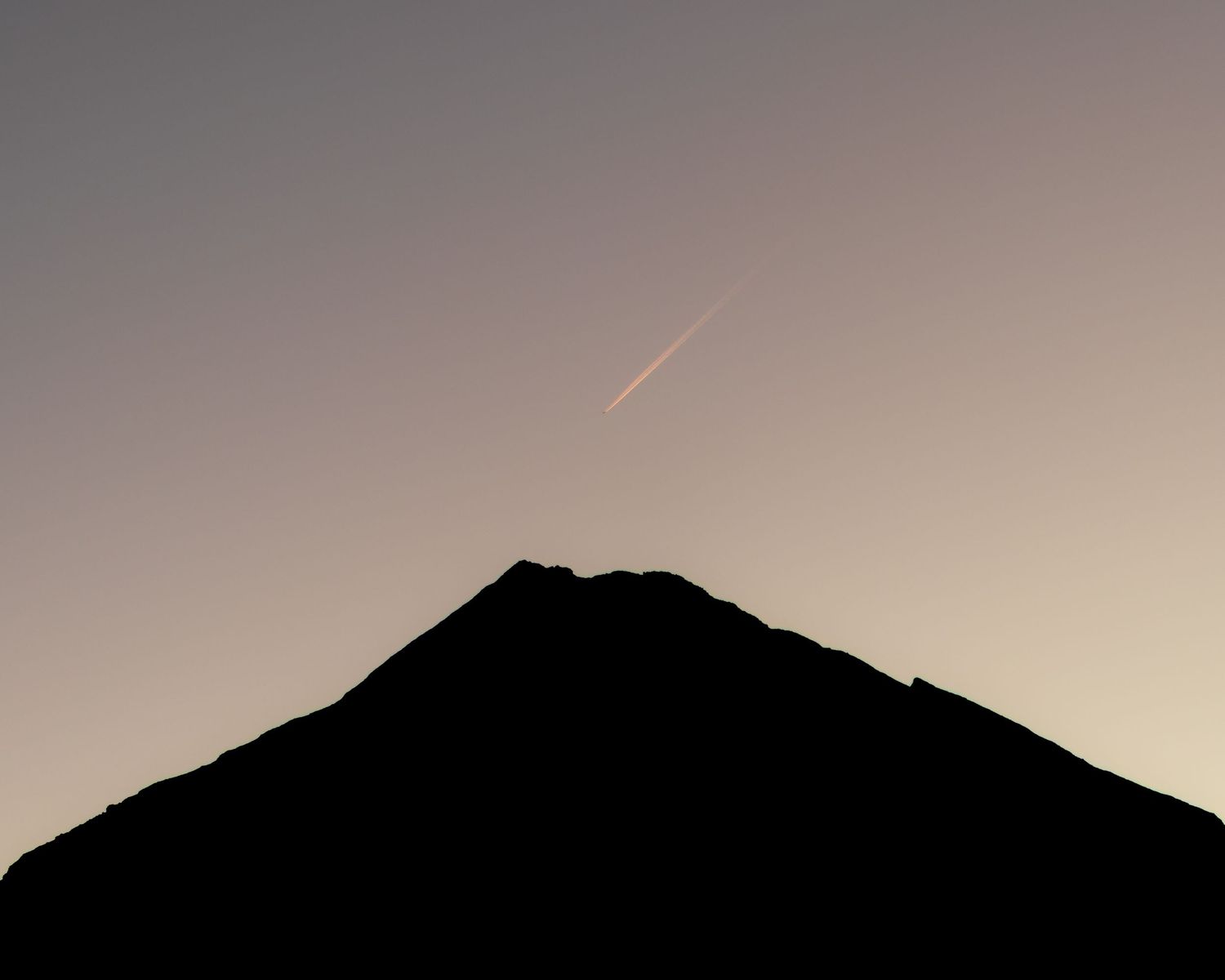 Mount Taranaki forms a dark silhouette against the fading sunset sky in Taranaki, New Zealand, with a faint aircraft trail overhead.