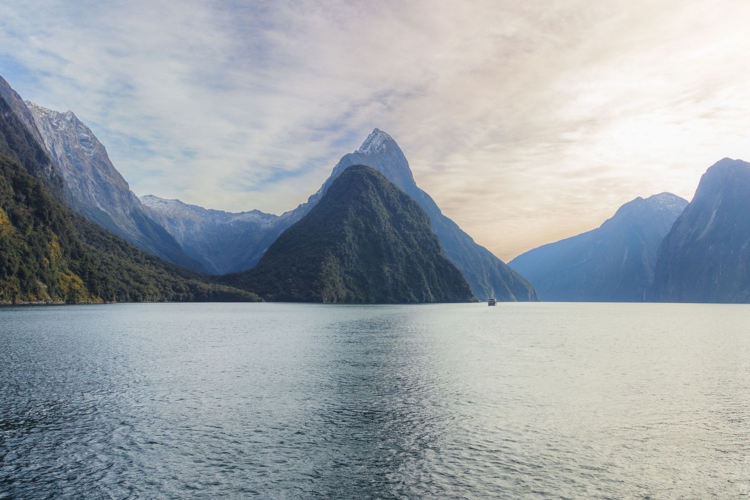 Mountain peaks rise above the calm waters of Milford Sound in Fiordland, New Zealand, beneath a bright sky.