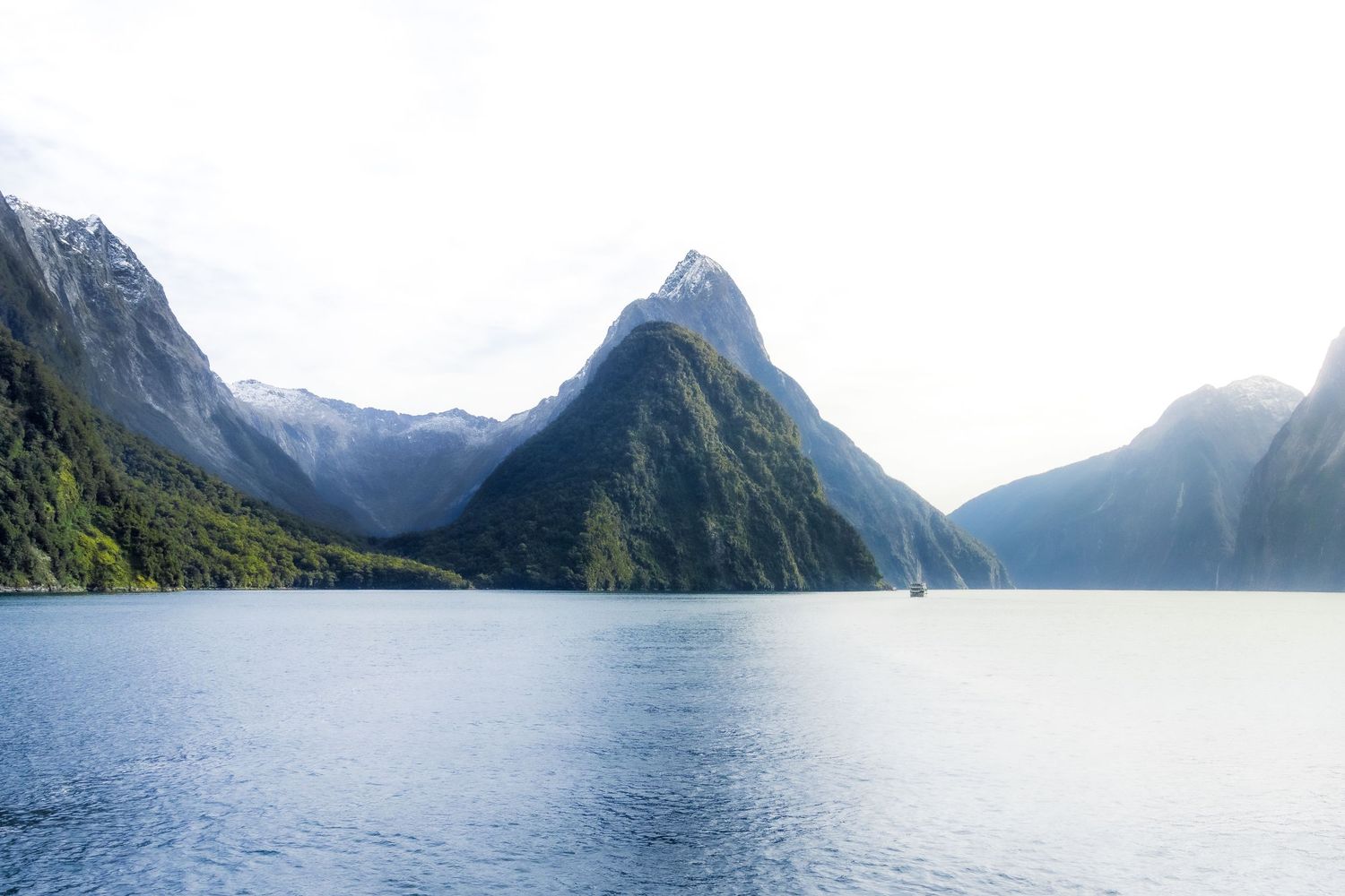 Mountain peaks rise above the calm waters of Milford Sound in Fiordland, New Zealand, beneath a bright sky.