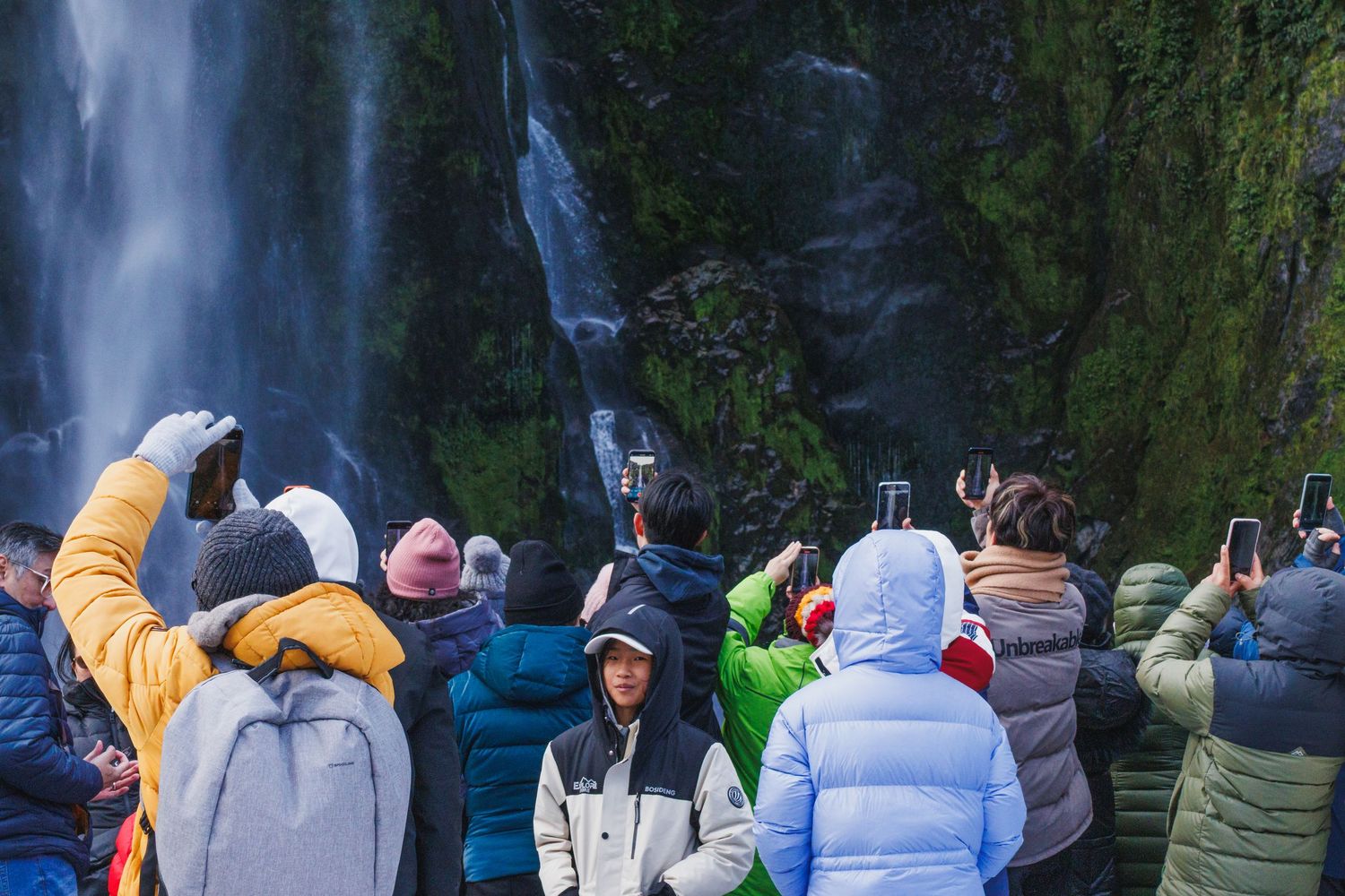 A crowd of visitors photographs a waterfall in Milford Sound, with one person facing back toward the camera.