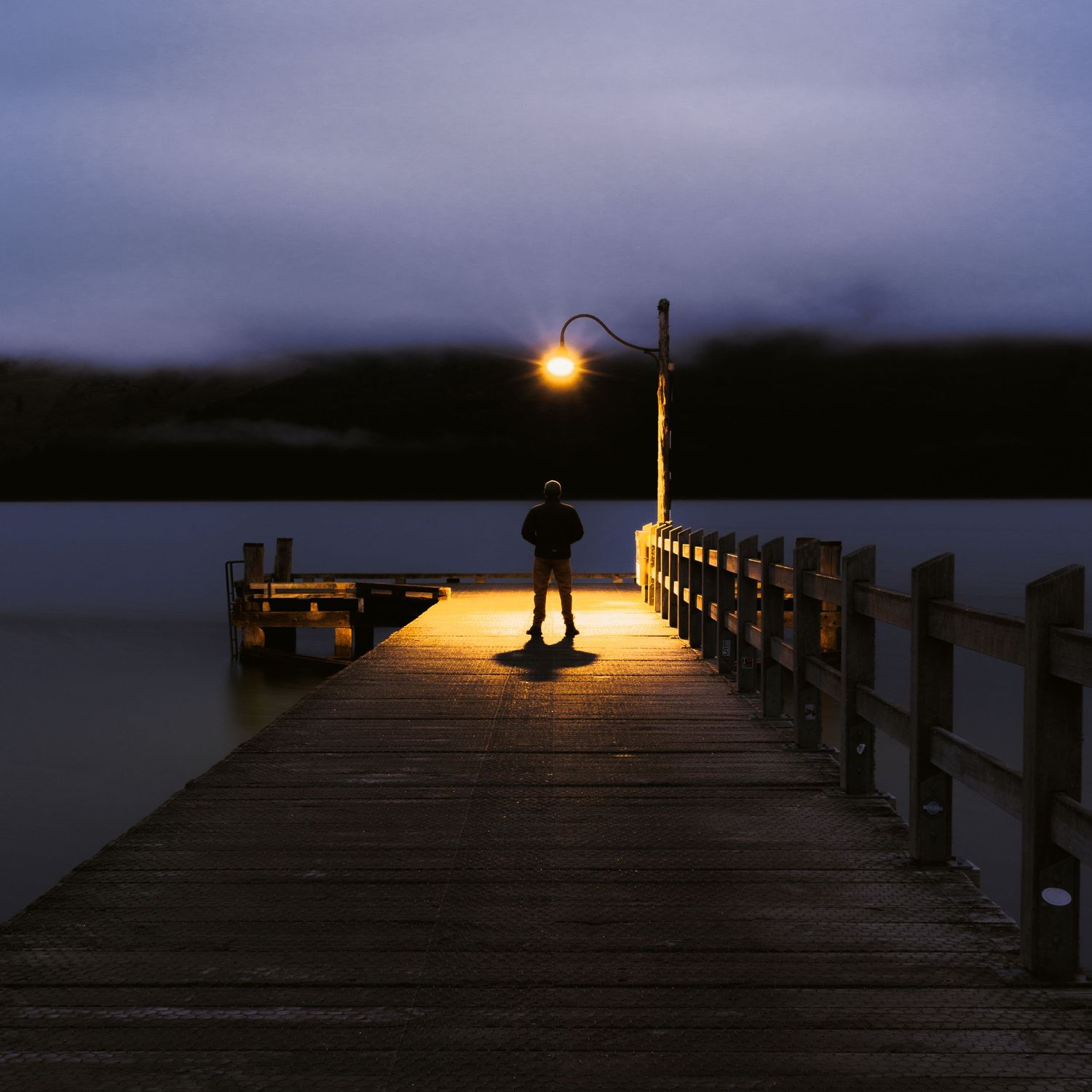 A lone man stands beneath a glowing lamp at the end of Glenorchy Wharf beside still Lake Wakatipu.