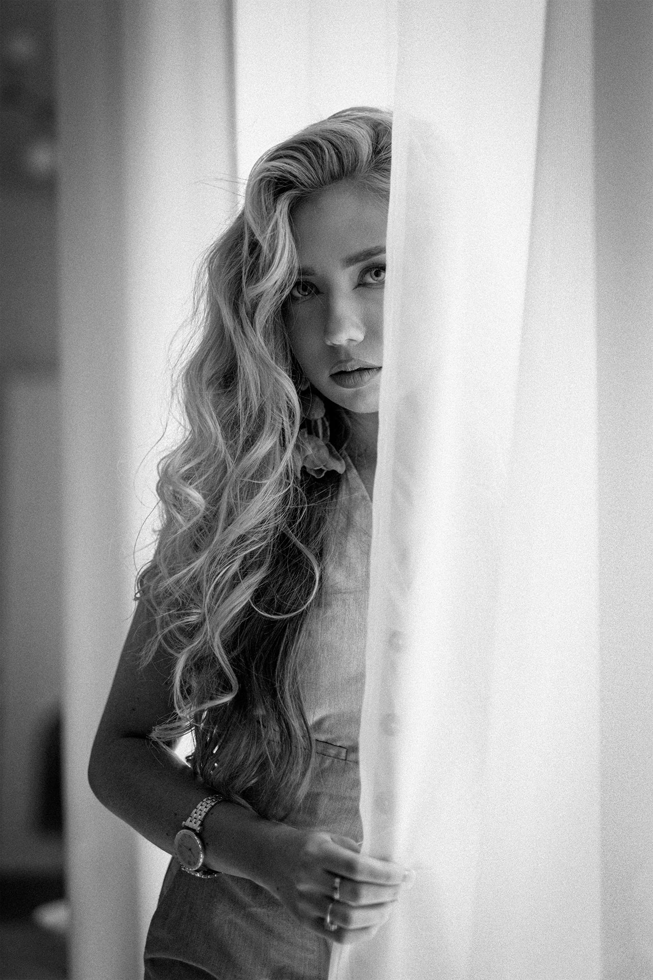 Black and white portrait of a woman looking through sheer curtains. Fine art women's portrait photography by Martyna Osuchowska, Częstochowa