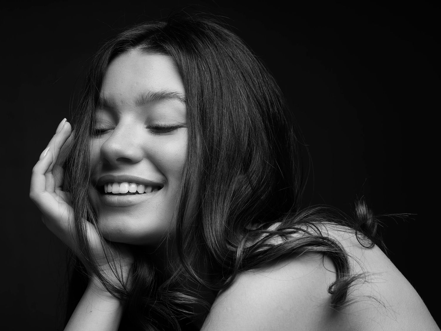Woman smiling joyfully with eyes closed, hand on cheek, in black and white portrait photo.