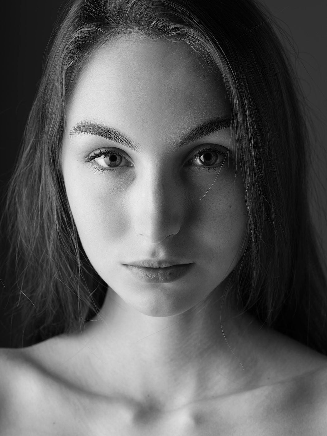 Black and white portrait of a young woman with long hair gazing directly at the camera against a dark background.