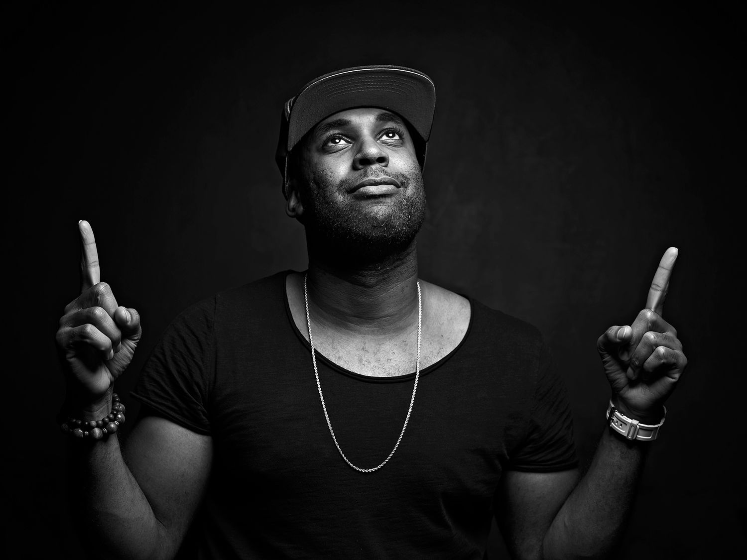 Black and white portrait of a young man in a cap and chain, pointing upward with both index fingers. John Davis
