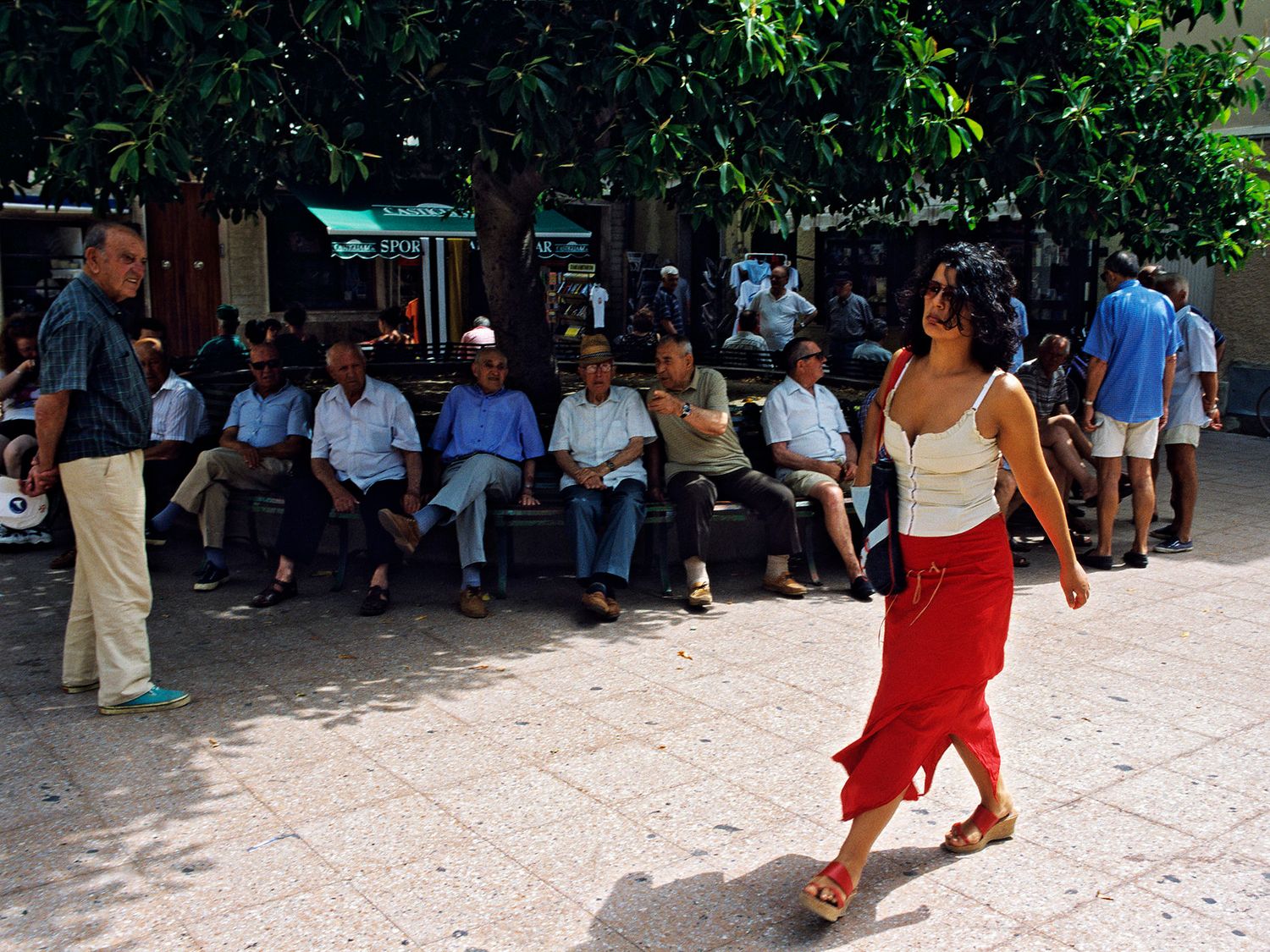 Woman in red skirt walking through a sunny Mediterranean plaza with men relaxing on benches under trees.