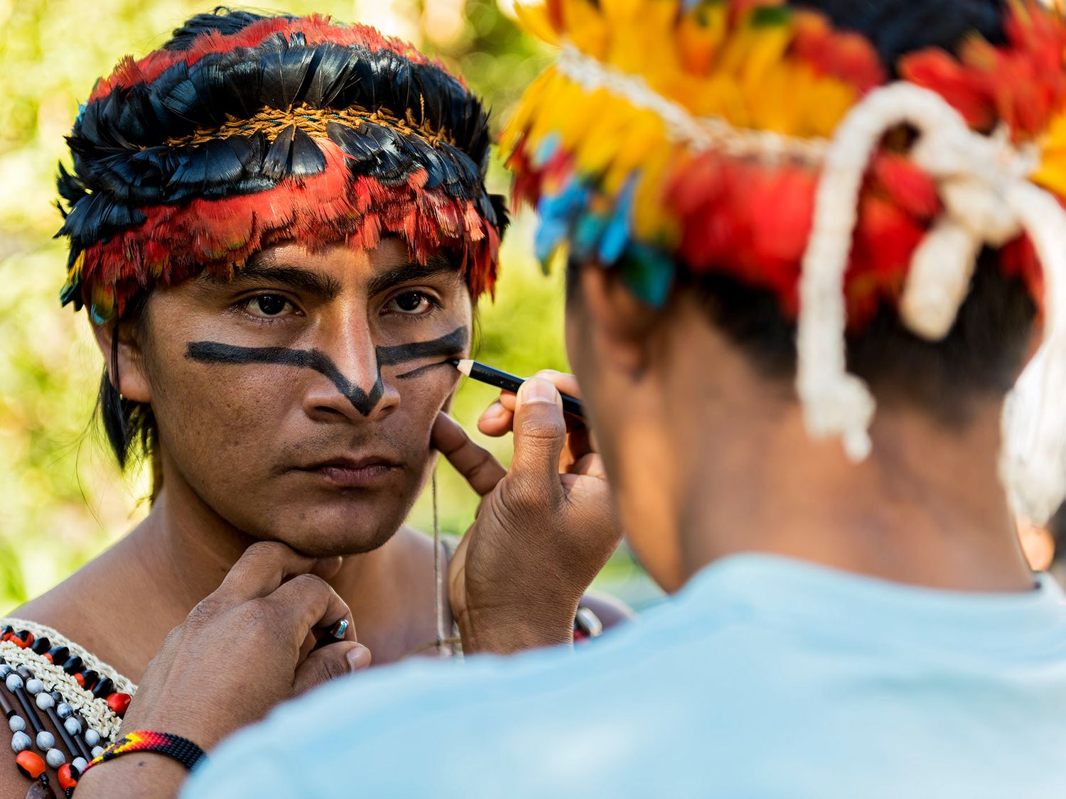 Indigenous man in feathered headdress gets traditional face paint applied during a cultural ceremony.