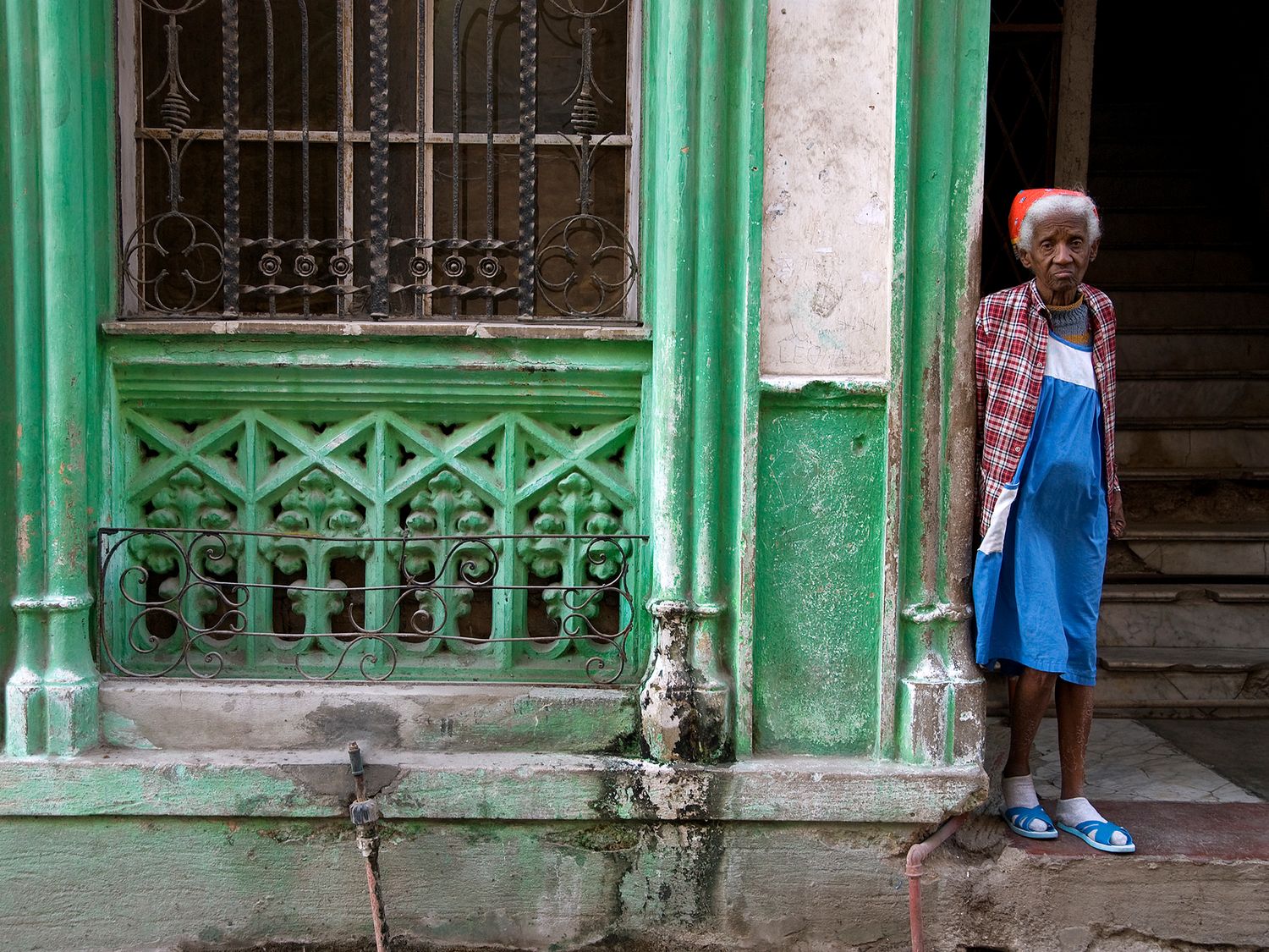 Elderly woman in blue apron standing beside a weathered green building with ornate iron window bars.