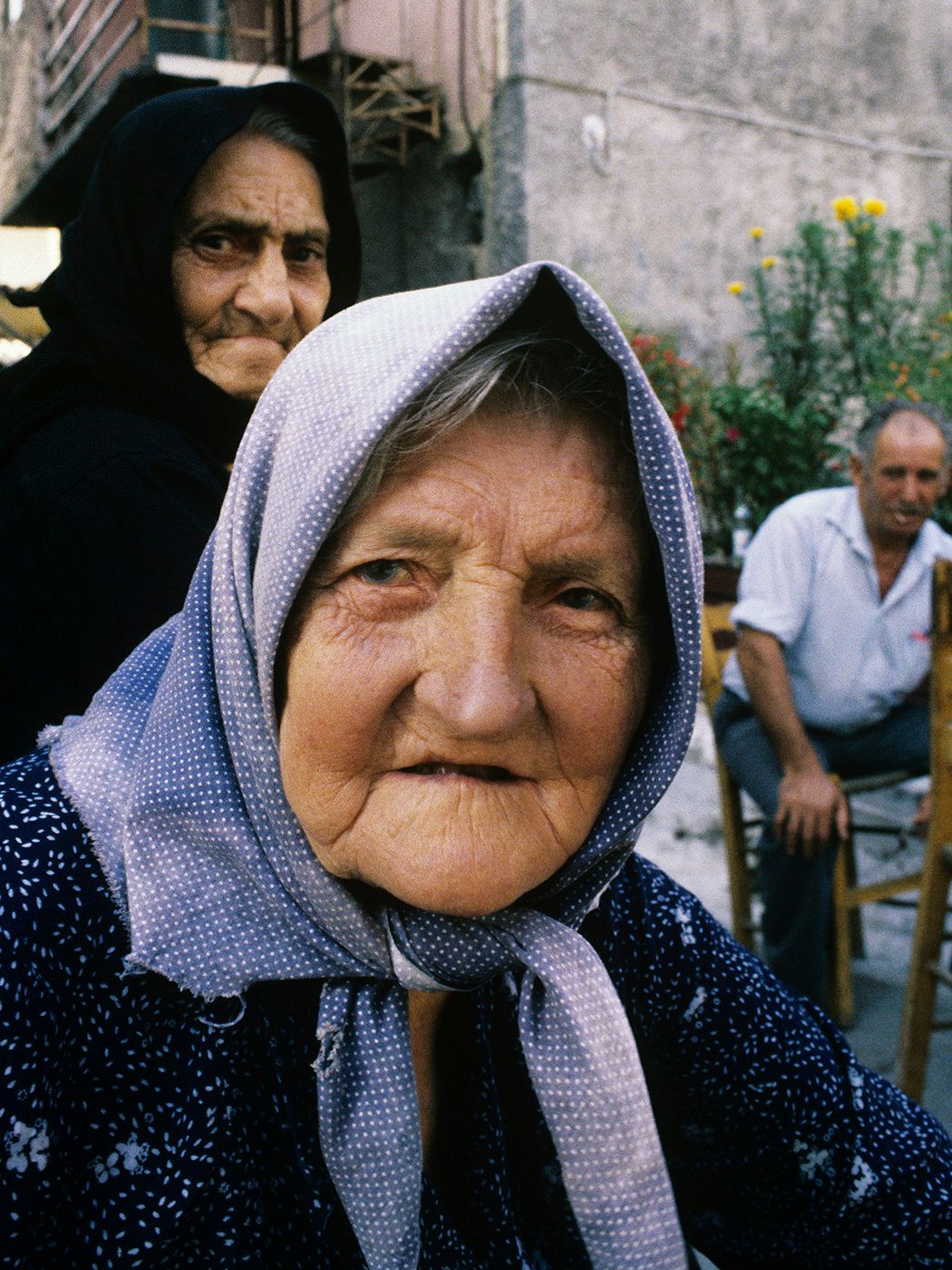 Elderly woman in a light blue headscarf with wrinkled face, another woman and man visible in background.