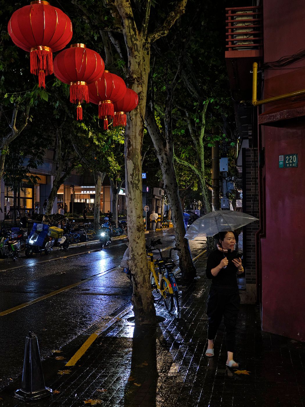 Woman with umbrella stands on rainy night street adorned with red Chinese lanterns, checking her phone beside a tree.